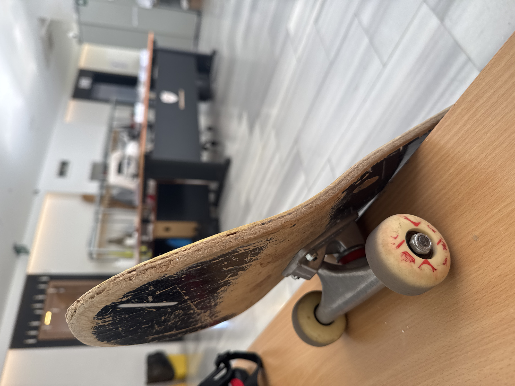 A close-up, medium-shot, angled photograph depicts a well-used skateboard resting on a light-colored, possibly wooden, surface. The skateboard deck is worn, with visible scratches and damage to the black graphic, and frayed edges. The wheels are cream-colored with red detailing and show signs of wear. The trucks appear to be silver metal.
The background is blurred, showing a brightly lit interior space with a tile floor and various pieces of furniture including a dark table or counter, and office equipment.
It is impossible to determine the exact location in Budva, Montenegro or any interactions in the environment from the provided image.