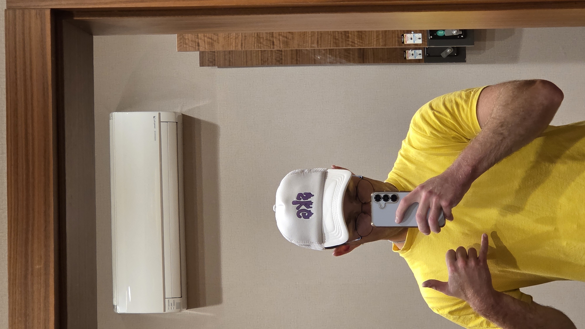 A person wearing a white trucker hat with purple drip-style lettering, a yellow t-shirt, and glasses is taking a selfie in a mirrored room. They are holding a silver smartphone in their right hand, obscuring their face, and making a shaka hand gesture with their left hand. In the background, an off-white air conditioning unit is mounted high on a plain, taupe-colored wall. To the right, a wooden framed mirror with vertical wood paneling is visible. The environment appears to be an indoor, possibly a changing room or bathroom, with a sense of clean, modern design. The location in the city of Bania, Bulgaria cannot be determined from the visual information provided.