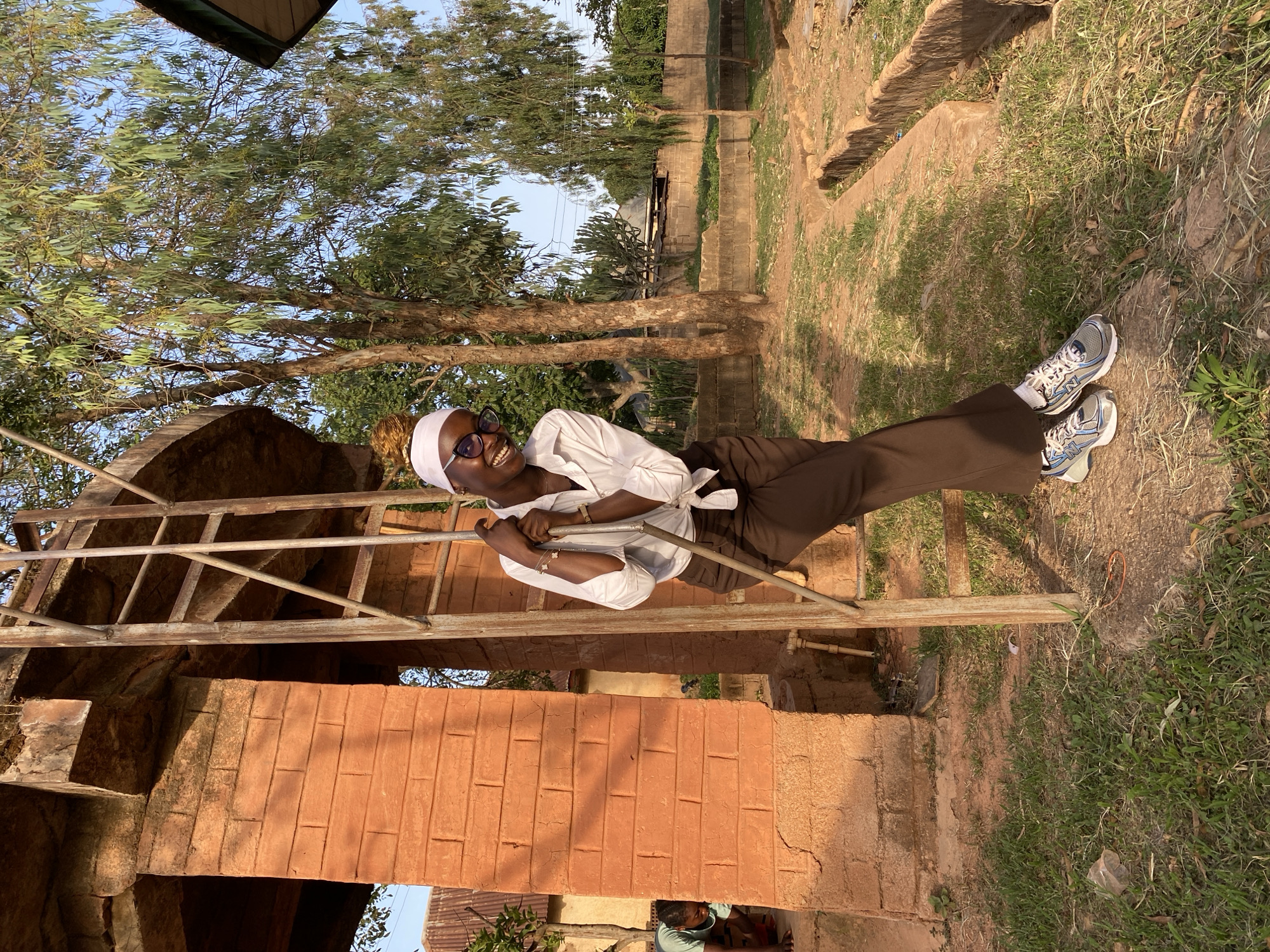 The image shows a woman in Vom, Nigeria, wearing brown pants, a white shirt, a white headscarf, and eyeglasses. She is smiling and clinging to a metal structure that appears to be a ladder or scaffolding. The structure is connected to a building constructed of red brick or clay. The surrounding environment shows grass, trees, and other buildings in the background. The location appears to be outdoors on a sunny day.