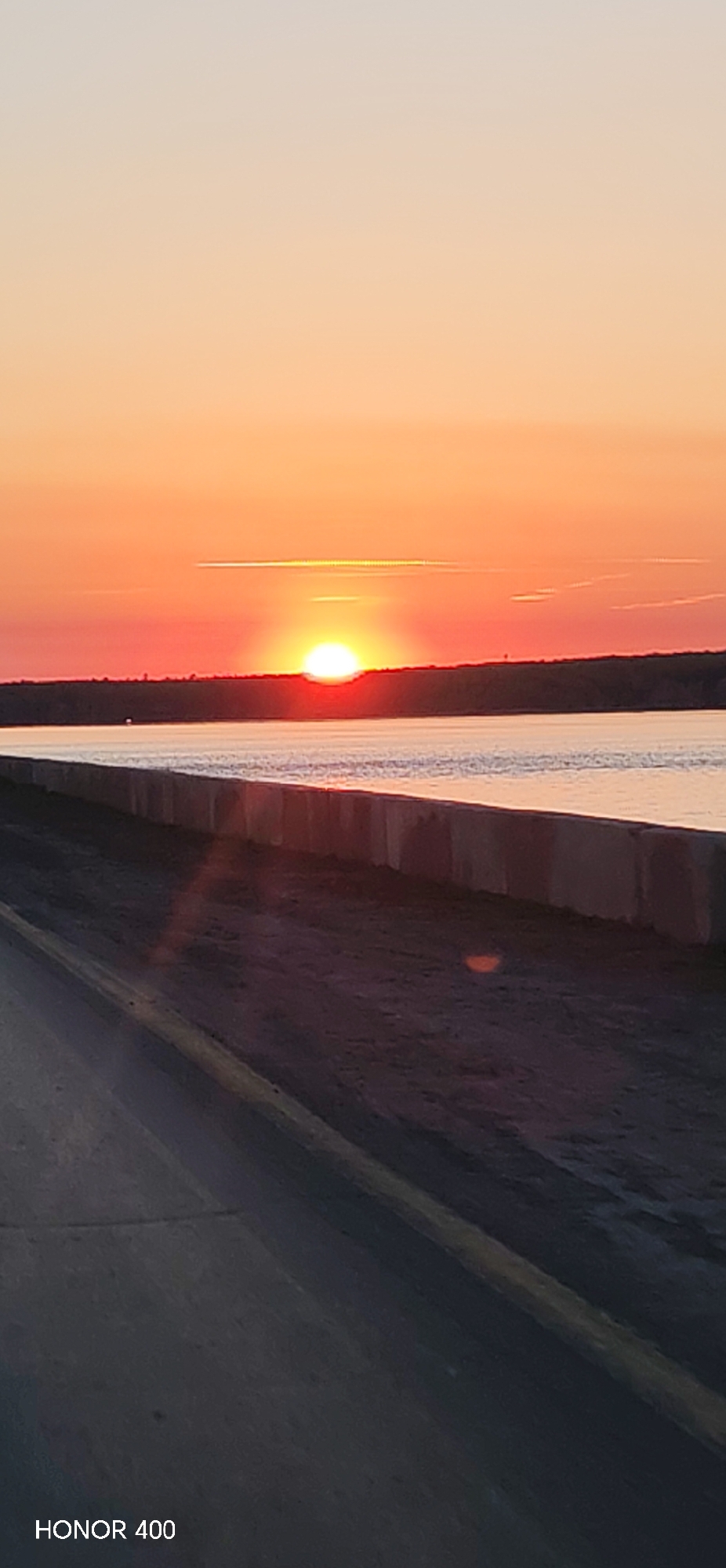 The image depicts a sunset over the Kama River in Chaikovsky, Russia. The sun is setting on the horizon, casting an orange and red glow across the sky. A concrete embankment runs along the riverbank. The foreground features a road, partially obscured by shadow. The camera is an HONOR 400, evidenced by the text in the bottom left corner.