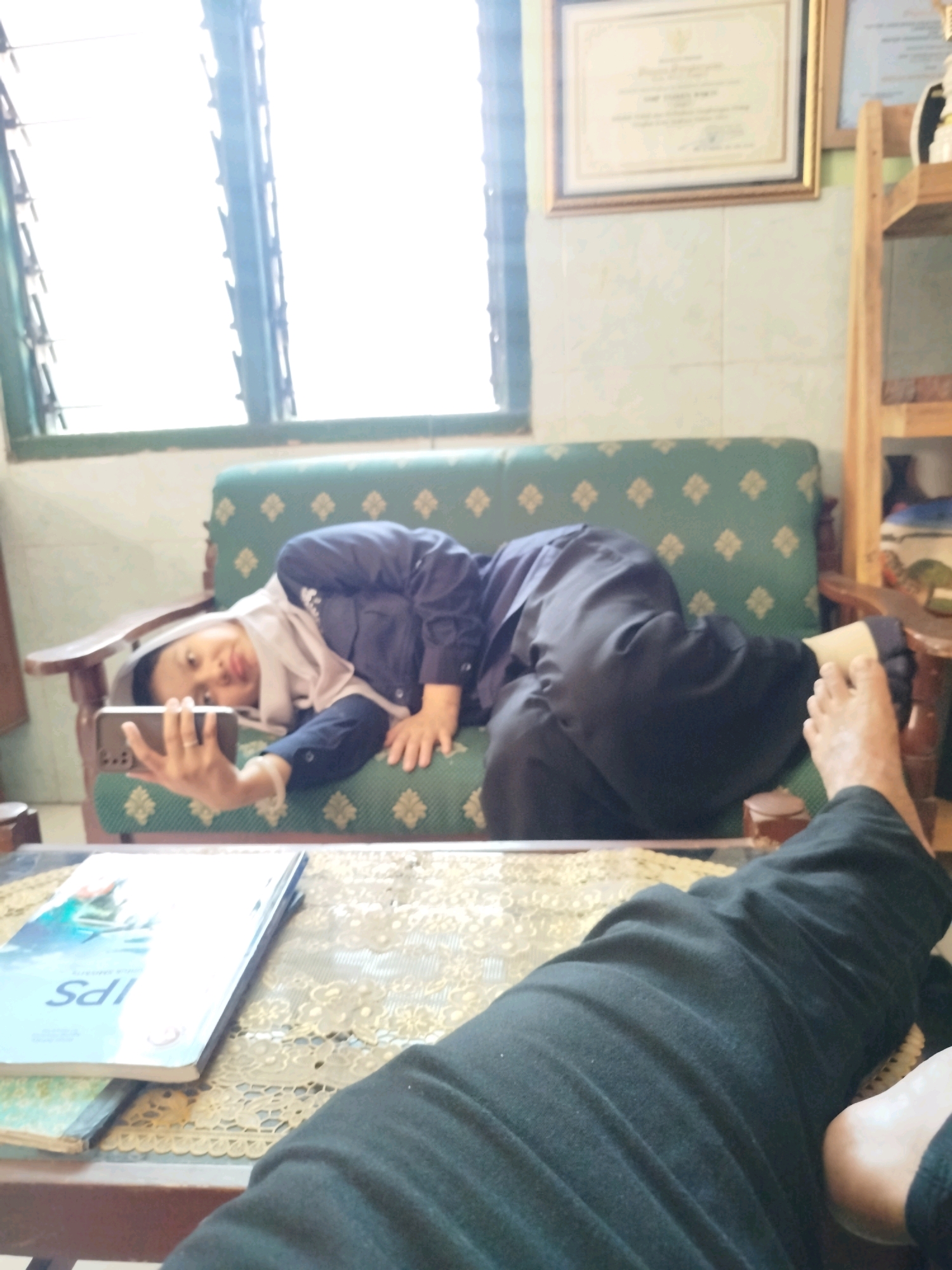 A person wearing a navy blue jacket and a light-colored hijab is lying on a green sofa with a diamond pattern, holding a smartphone in their left hand and taking a selfie. The person's right leg is bent, and their foot is visible in the foreground. In the background, a window with louvers lets in bright light, and a framed certificate hangs on the wall. A wooden shelf with items is also visible. A table with books is in front of the sofa. The scene appears to be an indoor setting in the city of Madiun, Indonesia, likely a home or office.