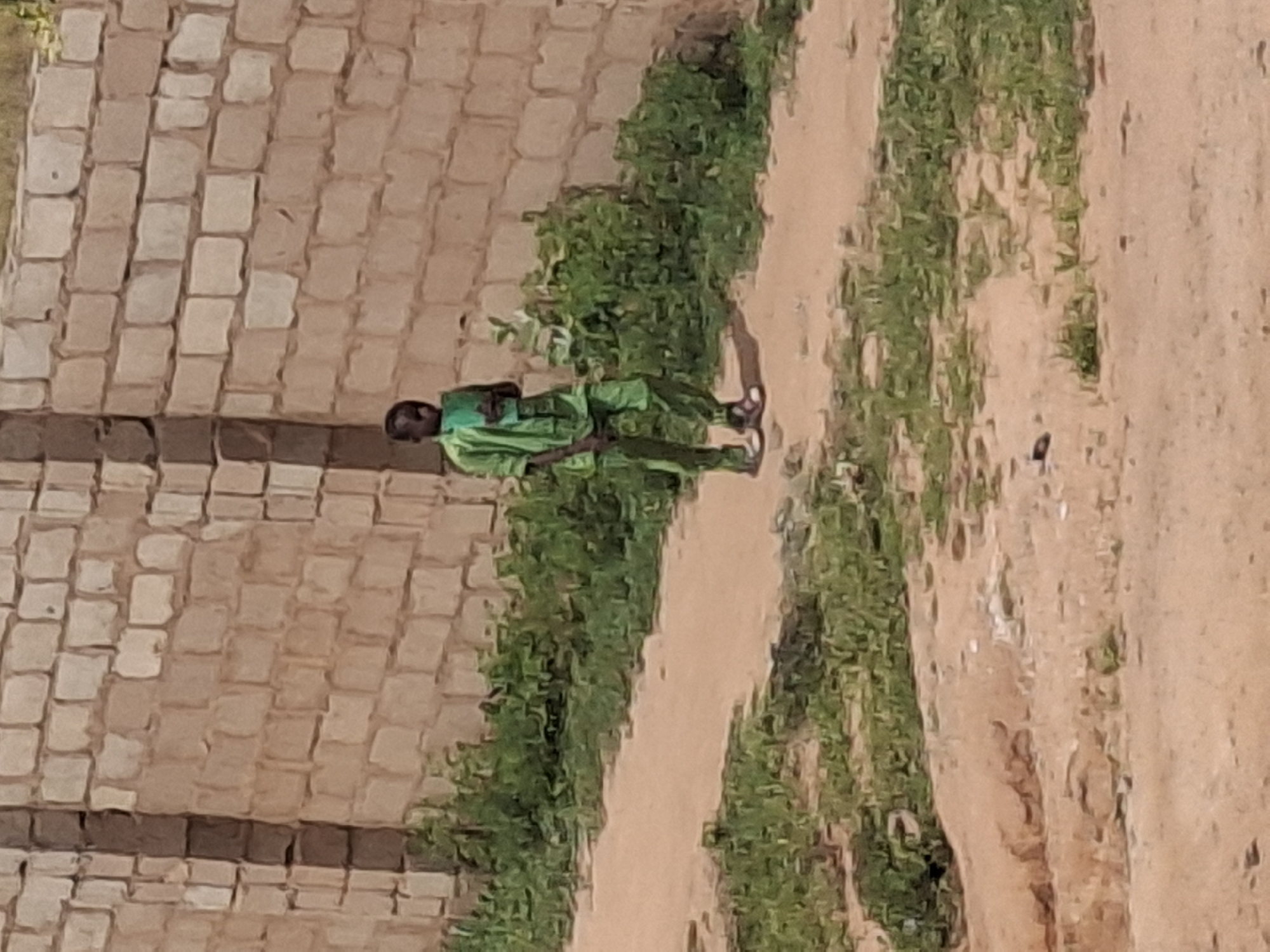 A young boy, appearing to be of African descent, stands on a dirt path in front of a brick wall. He is wearing a bright green, patterned traditional outfit with matching pants and a long-sleeved shirt. The boy is looking downwards at something he is holding in his hands. The environment appears to be outdoors, with sparse green vegetation growing along the path and at the base of the wall. The wall is constructed of light-colored bricks with darker mortar. The lighting suggests it is daytime. The image was captured in Tumfure, Nigeria.