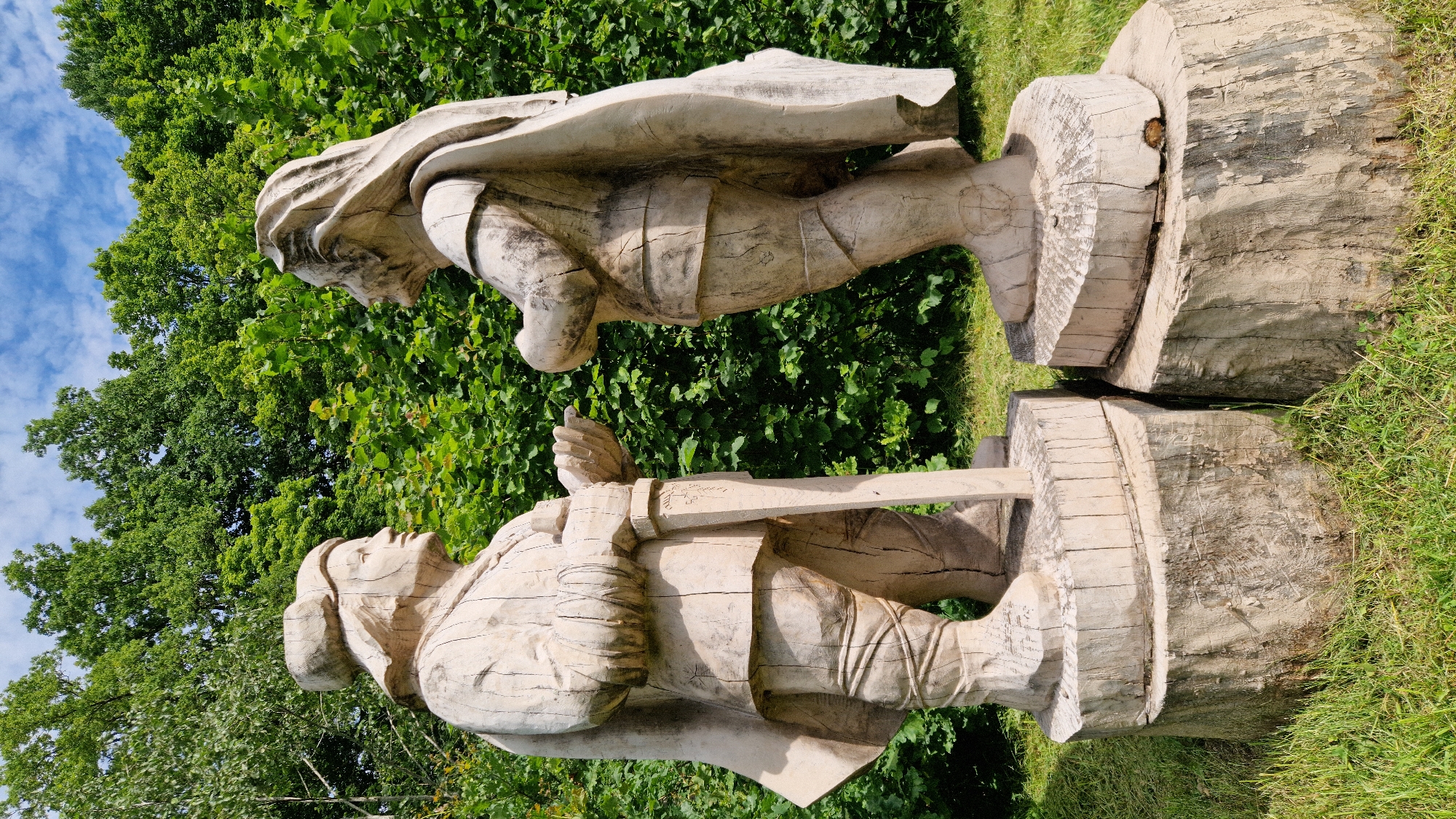 In Embūte, Latvia, two large, weathered wooden sculptures of figures stand side-by-side on tree stumps. The sculptures, appearing to be of men in historical attire, are positioned facing each other with their hands outstretched as if in conversation. The figure on the left wears a cap and cloak, holding a sword upright between its legs. The figure on the right also wears a cloak and appears to have long hair. Both figures exhibit carved details of clothing, including belts and boots. The backdrop consists of lush green foliage and trees under a partially cloudy blue sky. The foreground shows green grass and the rough texture of the wooden bases.