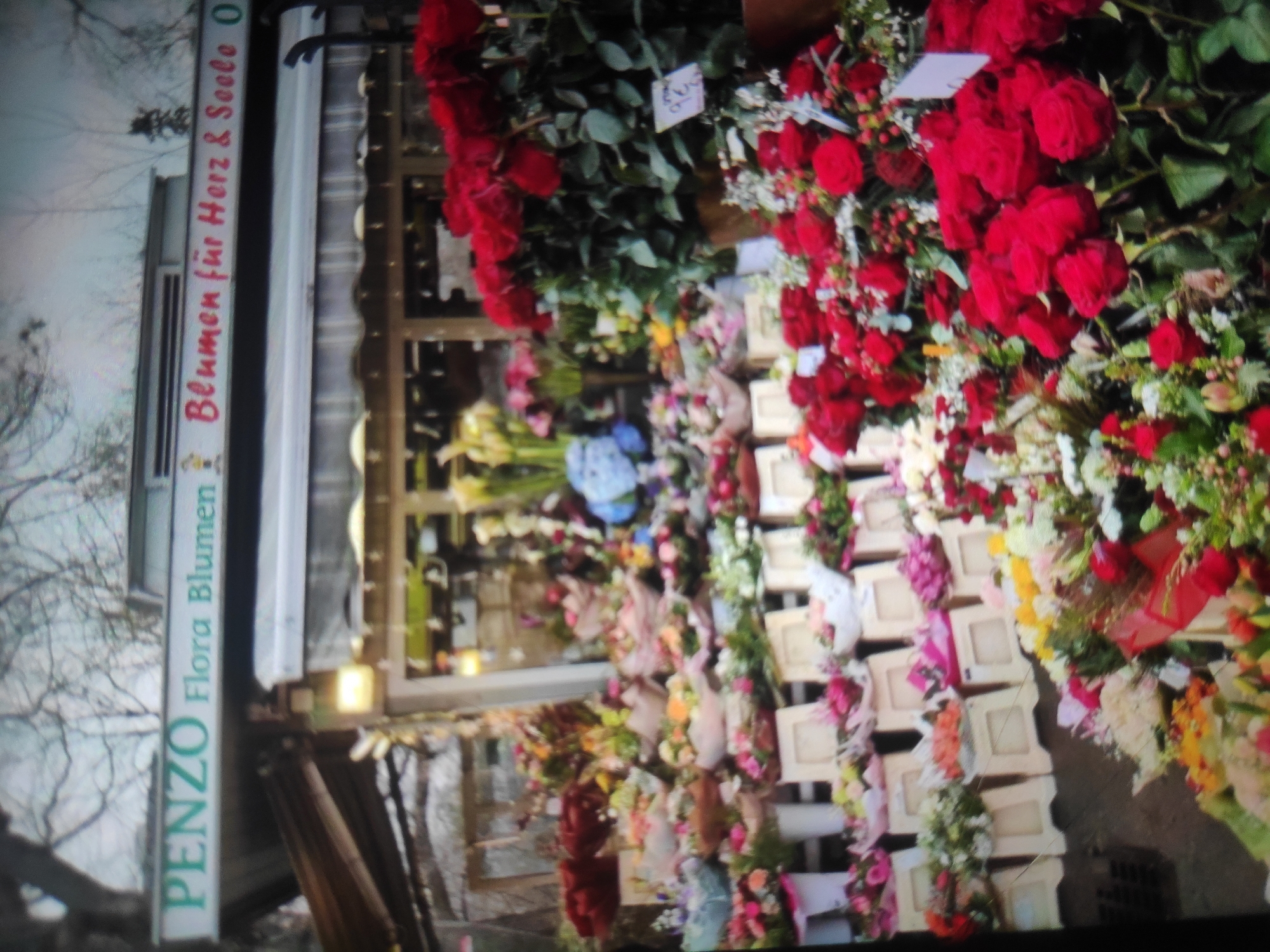 This is a close-up, slightly blurry, eye-level shot of a flower stall in Ceporan, Indonesia, displaying a vibrant assortment of flowers. The signage above the stall reads "PENZO Flora Blumen Blumen für Herz & Seele". The stall is packed with various floral arrangements, including numerous red roses in the foreground, bunches of pink and white flowers, and some yellow and blue flowers further back. The flowers are presented in baskets and wrappings, suggesting they are for sale. The background shows the covered structure of the stall with some hints of what appear to be buildings or trees. The overall impression is of a bustling, colorful flower market.