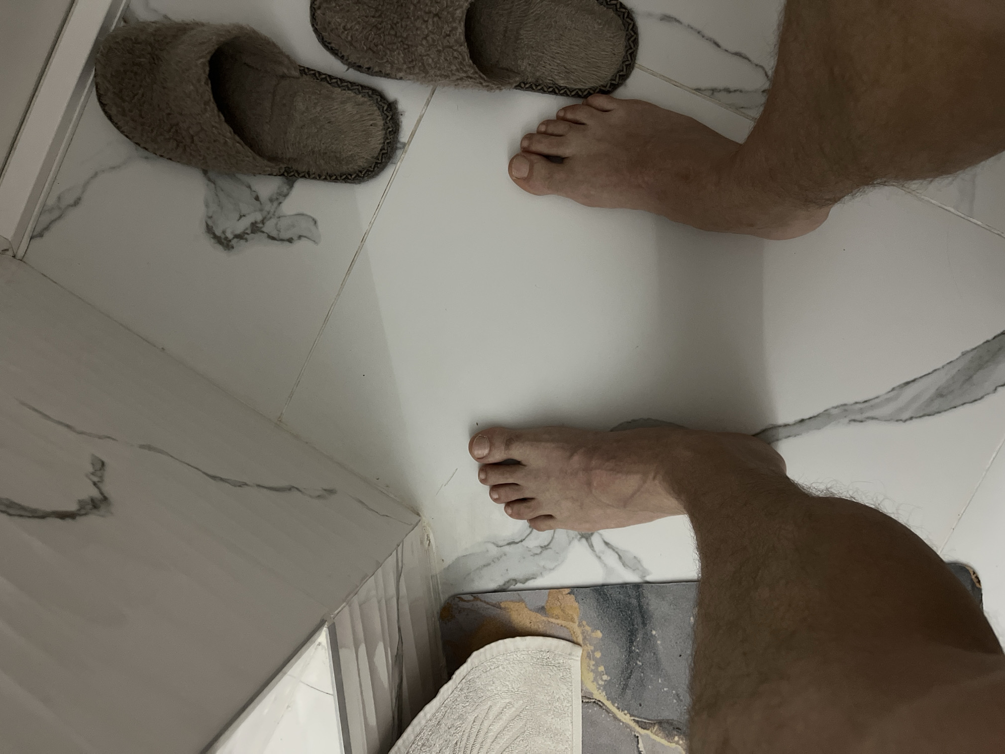 An overhead shot captures a person's bare feet and lower legs on a clean, white tiled floor featuring subtle grey marble-like patterns. Two well-worn, furry brown slippers are visible in the upper left portion of the frame, positioned near the person's feet. The person's legs are hairy.
In the bottom right corner, a segment of a bath mat or rug with an abstract grey, white, and gold design is partially visible, along with a folded white towel. A white rectangular structure, possibly part of a shower frame or bathroom fixture, runs along the left edge of the image. The scene depicts an indoor setting, strongly suggesting a bathroom or dressing area in Sochi, Russia. No specific activity is taking place beyond the person standing on the floor, and no text is visible. The time of day and any emotions cannot be determined from the image.