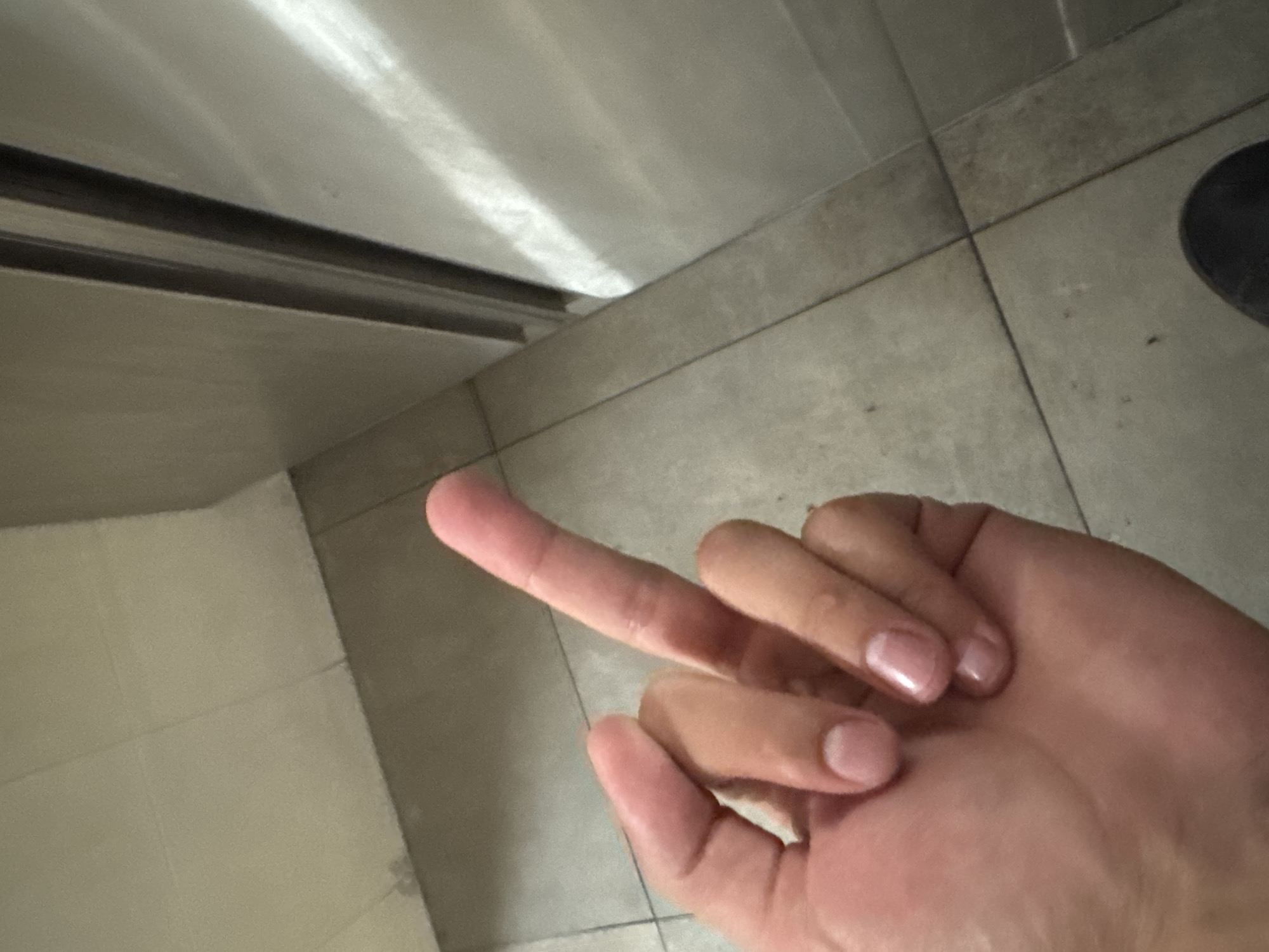 A hand, positioned with the middle finger extended, is in focus against a background consisting of tile flooring and a stainless steel surface. The tiles are a neutral gray color, while the stainless steel surface appears reflective. The scene suggests an indoor setting, and the overall composition creates a close-up perspective. Location information is not discernible from the image.