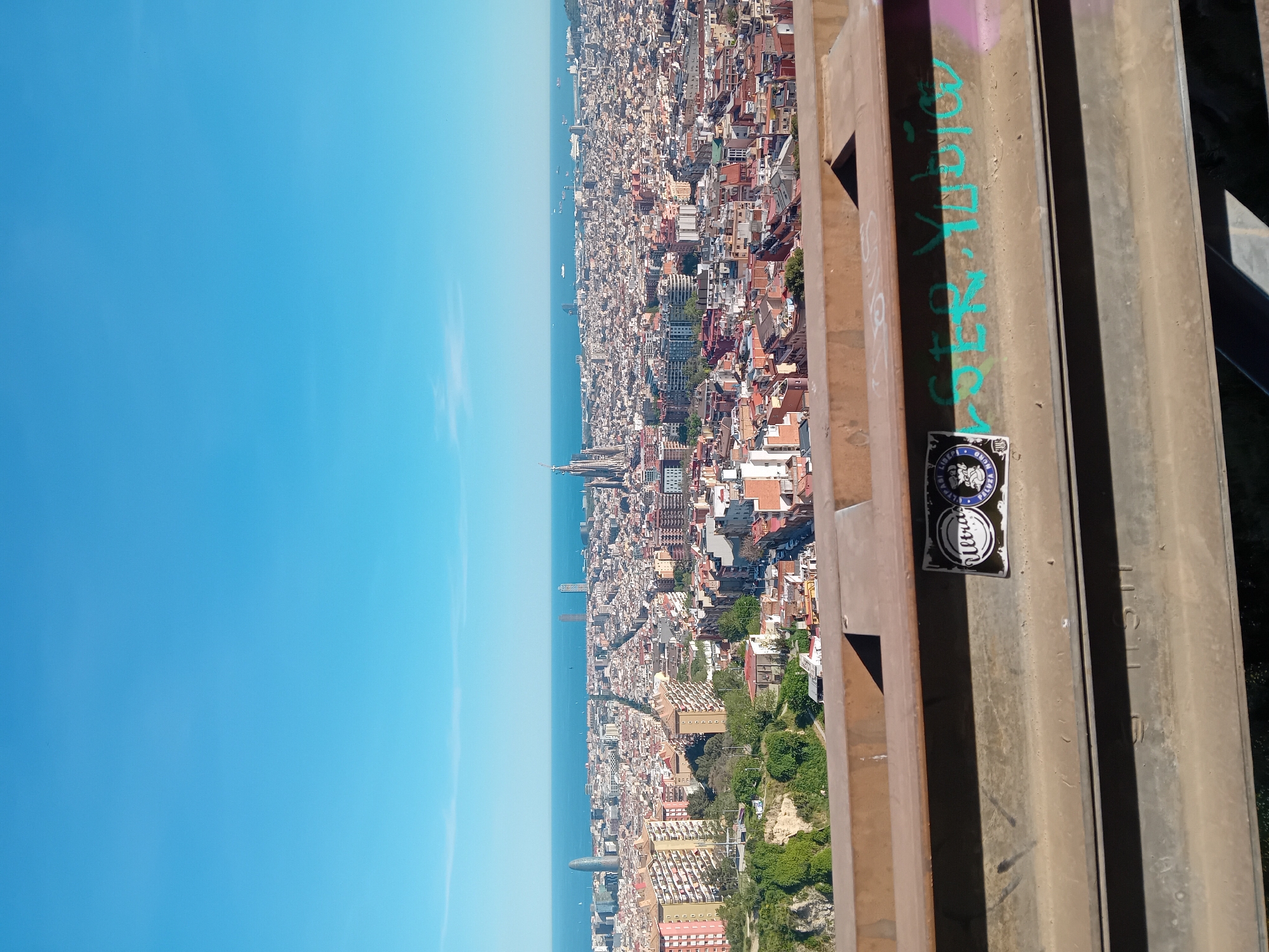 Here is a detailed description of the image:
The image shows a high-angle view of a coastal city, likely Barcelona, taken from a structure with metal beams. The sky is clear and blue, meeting the sea on the horizon. A dense urban landscape stretches along the coastline, with buildings of varying heights and colors, predominantly reddish-brown and beige. There are occasional green spaces, indicating parks or tree-lined streets.
In the foreground, the metal structure is visible, featuring graffiti and stickers. The graffiti is in teal or green paint, and the stickers appear to be circular and possibly branded. The metal beams have a worn, weathered appearance.