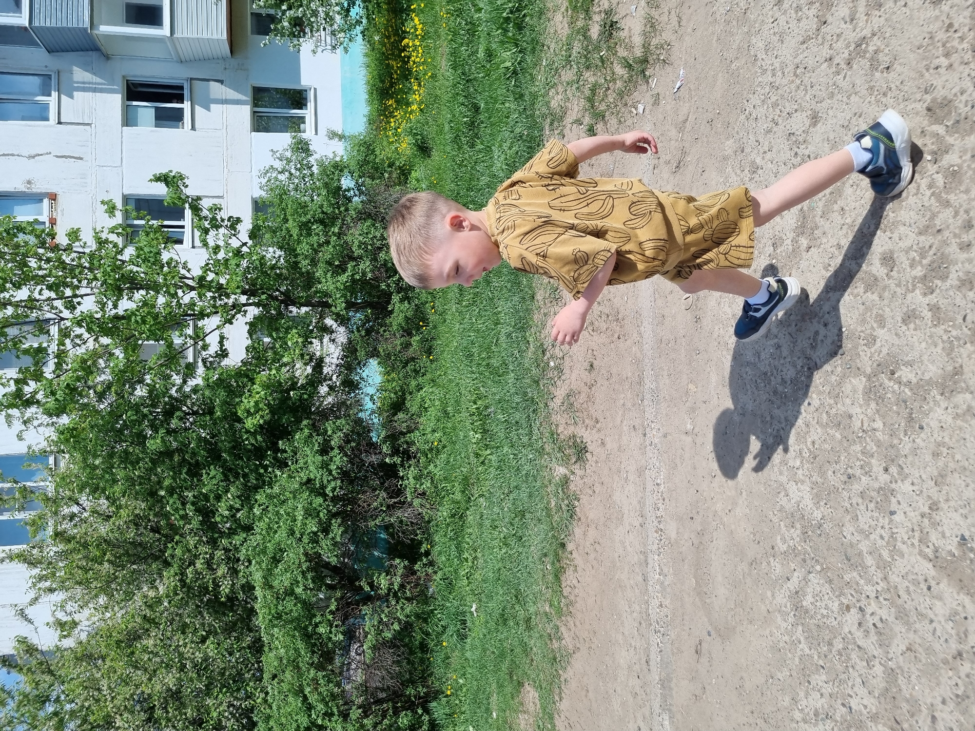 A photograph taken on a sunny day depicts a young child running along a dirt path. The child is wearing a yellow banana-print shirt and shorts with blue and white sneakers. The dirt path is edged by a patch of green grass. In the background is a light-colored apartment building, partially obscured by large green trees. The location is likely in a residential area of Chaikovsky, Russia, as indicated by the style of the apartment building. The child's shadow is cast on the dirt path in front of them.