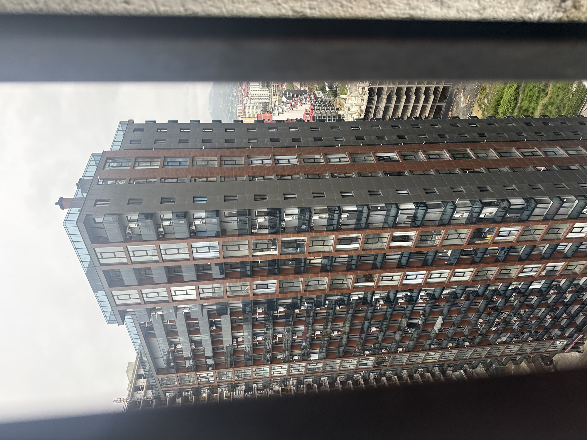 A tall, modern apartment building dominates the scene, viewed from an elevated position through what appears to be a window frame or balcony railing. The building has a geometric facade with alternating grey and brown horizontal bands, and numerous windows equipped with external air conditioning units. The top floors feature glass enclosures.
In the background, a cityscape unfolds, including buildings of varying heights and colors, a construction site with skeletal structures, and a patch of green vegetation. The overall atmosphere suggests the city of Batumi, Georgia. The sky is overcast, creating a diffused light. The photo has a slightly tilted perspective.