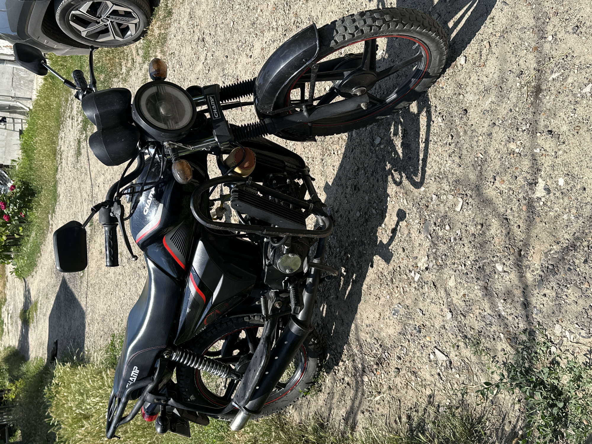A black motorcycle with red accents is parked on a gravel path outdoors. The motorcycle has a prominent round headlight, a black seat with red stitching, and a black exhaust pipe. The "CHAMP" logo is visible on the motorcycle. In the background, there is a dirt road leading away from the motorcycle, with grass and some foliage on either side. A dark-colored vehicle's wheel is visible to the right of the motorcycle. The scene appears to be in an outdoor, rural or semi-rural setting, possibly in the city of Comrat, Moldova, given the context. The lighting suggests it is daytime with bright sunlight casting shadows. There are no people visible in the image.