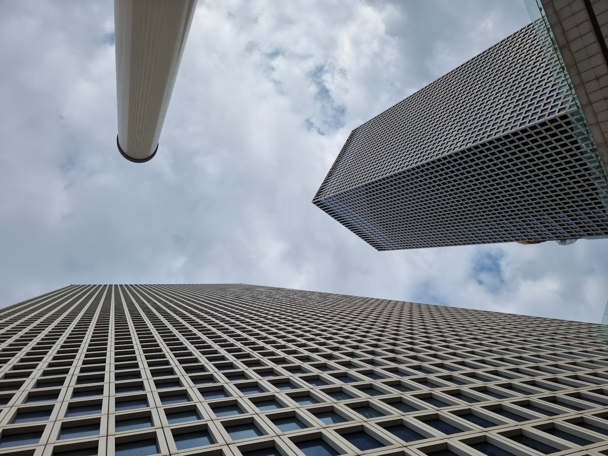 The image depicts a low-angle view of three architectural elements set against a cloudy sky. On the left, a cylindrical structure, possibly a column or a section of a building, is partially visible. To the right, a modern skyscraper with a densely patterned facade rises towards the sky. The facade is composed of a repetitive grid of squares, each appearing to frame a window. The largest building is at the bottom of the image, seemingly a skyscraper with many windows, the same style and structure as the skyscraper on the right, but in a closer view.
Based on the architectural styles of the buildings, particularly the large skyscraper in the foreground, and the information provided, the image is most likely taken in Tel Aviv, Israel. The buildings appear to be modern and situated in a business or commercial district.