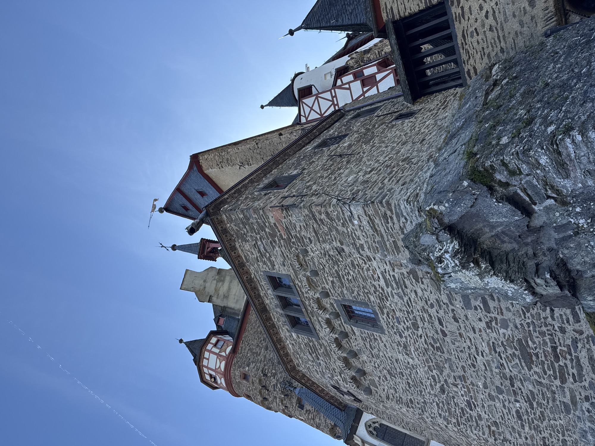 The image depicts Eltz Castle in Wierschem, Germany. The view is angled upwards, showcasing the stone walls, towers, and architectural details against a clear blue sky. The castle is built on a rocky outcrop, and parts of the natural rock formation are visible. There are various architectural elements, including windows, turrets, and decorative roofs. The castle exhibits a mix of colors, primarily stone grays, white, and red.