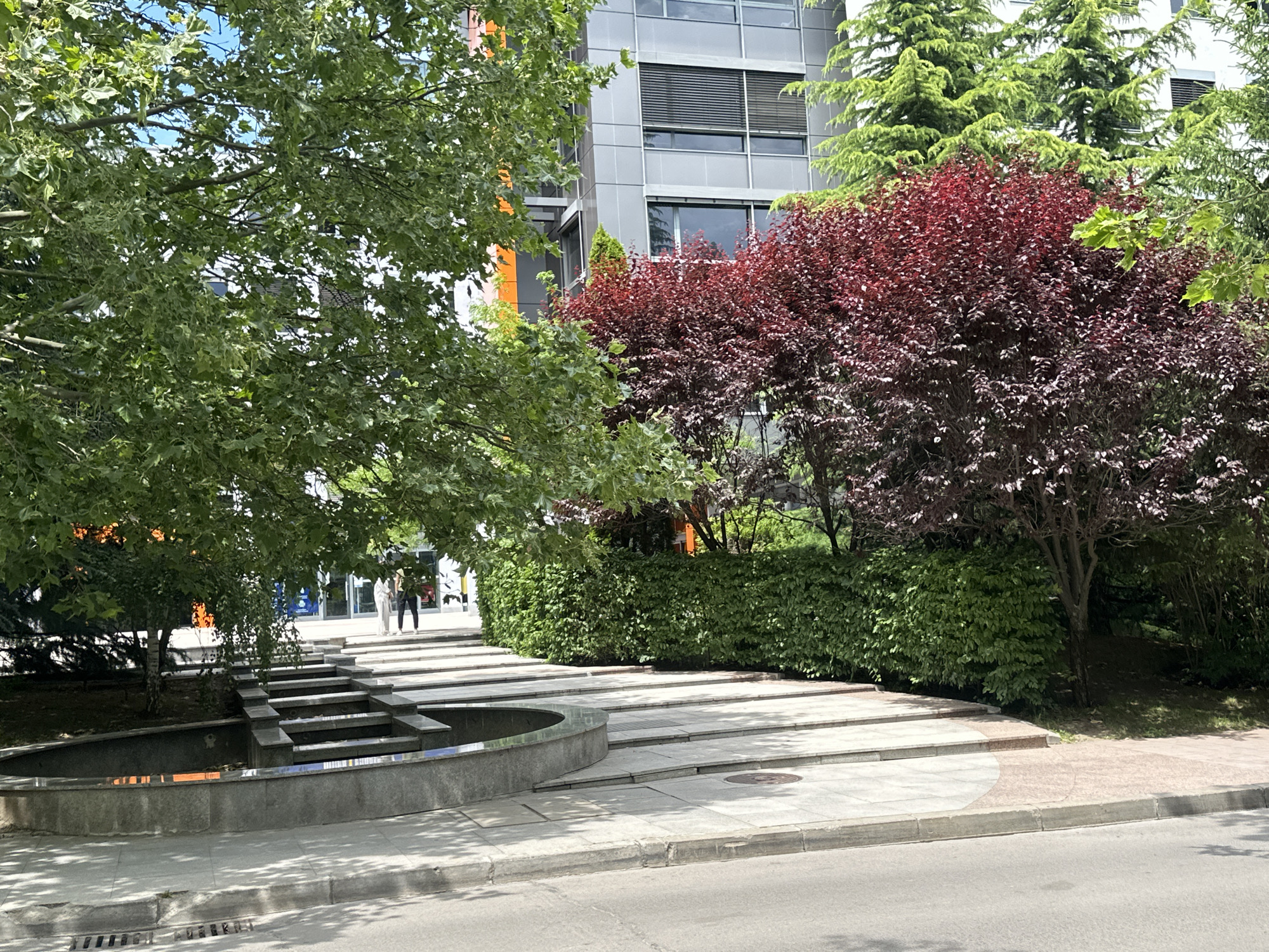 The image shows an outdoor scene in front of a modern building with a grey facade and orange accents. In the foreground, there is a paved area with a curved water feature and steps leading up. Lush green trees and bushes surround the area, with a prominent red-leafed tree adding a splash of color. Two people are visible walking up the steps in the middle ground. The scene appears to be set during the daytime, under what seems to be sunny weather, casting shadows on the pavement. The presence of a building with glass windows and modern design suggests an urban or business park setting. The overall impression is one of a well-maintained public space integrated with a contemporary architectural environment.