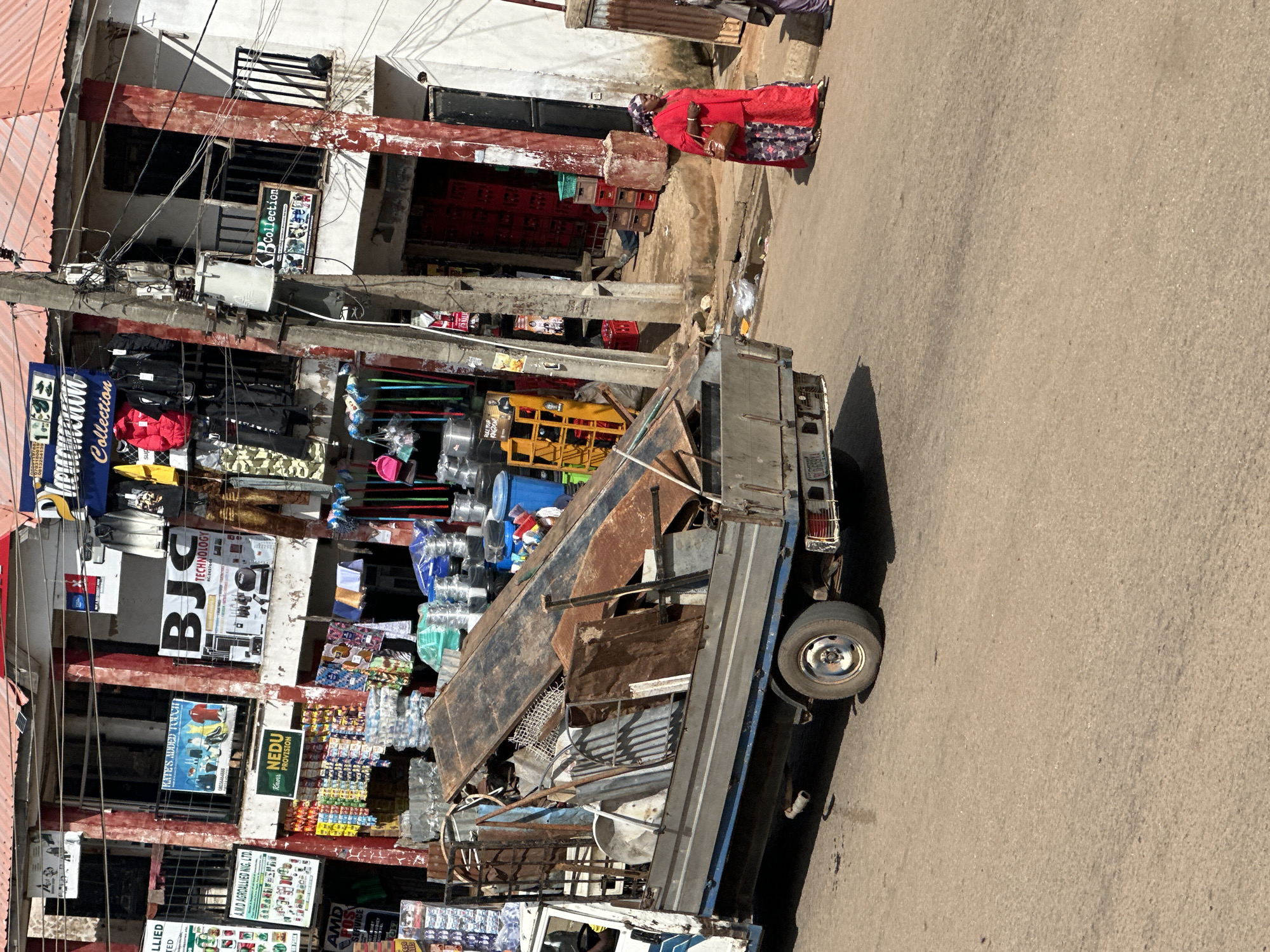 This image depicts a street scene in Bukuru, Nigeria, likely during the daytime given the bright sunlight casting shadows. The foreground is dominated by a utility truck loaded with what appears to be scrap metal and other materials, parked on a paved road.
The background reveals a bustling market or commercial area. On the left side of the image, a multi-story building with red trim and open storefronts is visible. These stores display various goods, including what seem to be textiles, housewares, and possibly electronics, indicated by signage like "KBcollection," "BJC TECHNOLOGY," and "Phenomenon Collection." There are also advertisements for "NEDU PROVISION" and "A.M.D AGROALLIED NIG. LTD."
A woman in a vibrant red garment is seen walking on the right side of the road, carrying a brown bag. Her attire and the surrounding environment suggest a cultural context specific to the region. The presence of overhead wires and the architecture of the buildings are typical of urban or semi-urban settings in Nigeria. The overall impression is one of a lively commercial hub.