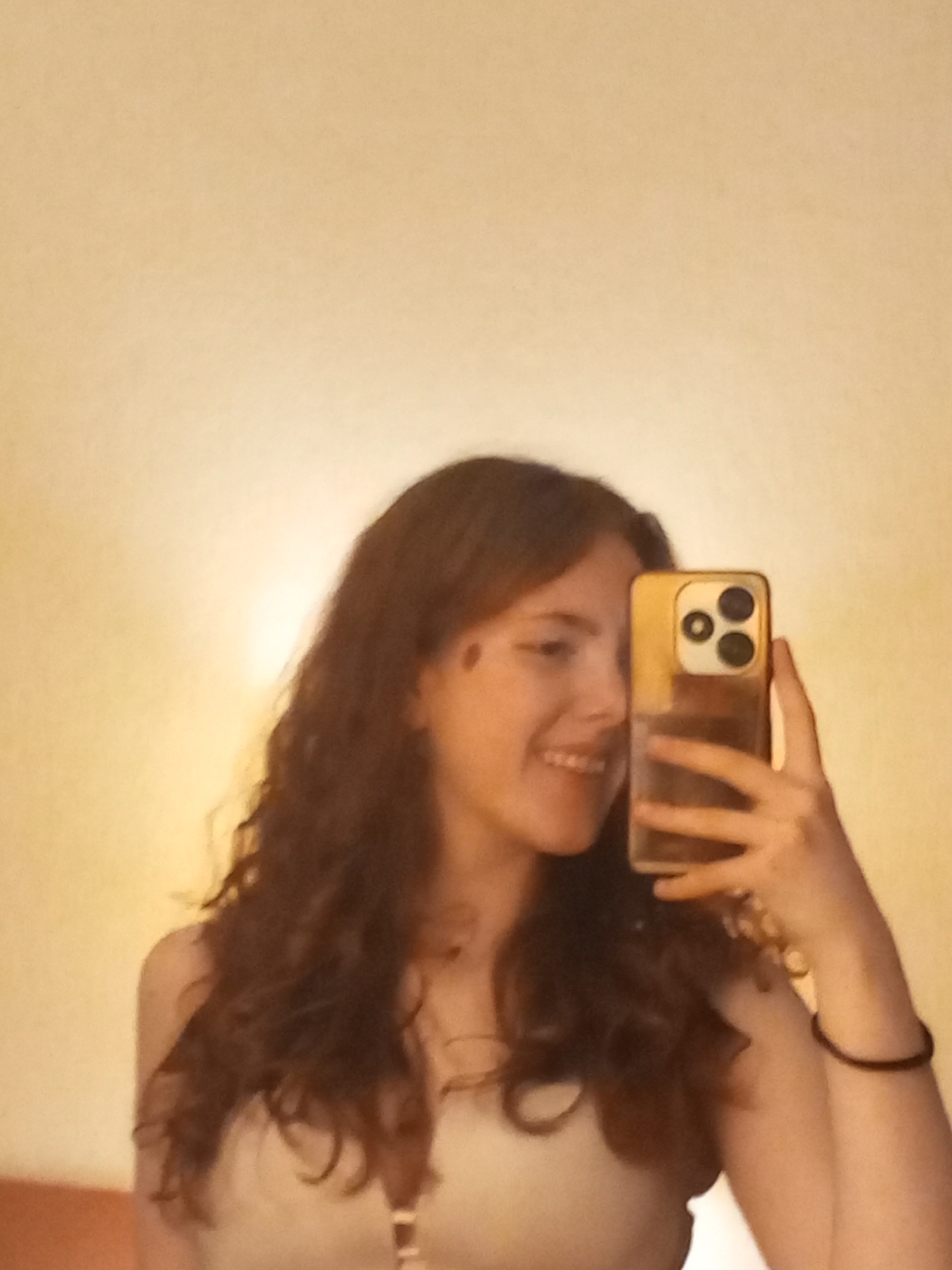 A young woman is taking a selfie indoors. She is holding a smartphone with a gold-colored case up to her face, partially obscuring it. She is smiling slightly. She has long, curly, dark hair. She is wearing a light-colored top with thin straps. There is a black bracelet on her left wrist. The background is a plain, light-colored wall. The location is not discernible to the city of Saint Petersburg, Russia.