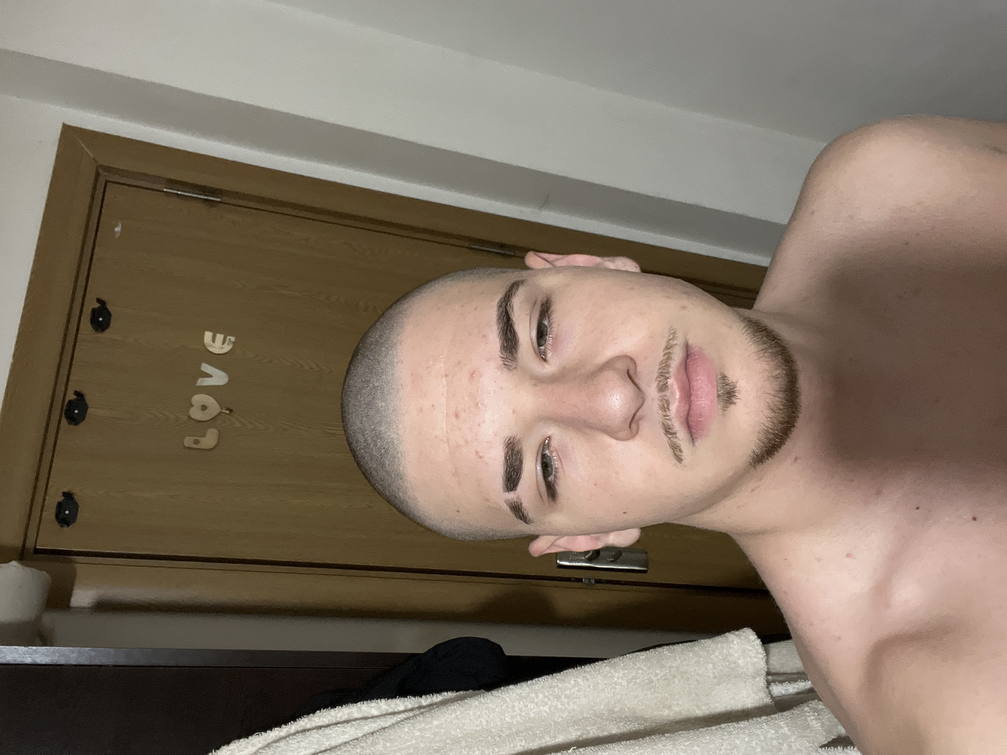 A close-up, rotated portrait shows a young man indoors, likely in Varna, Bulgaria. He is seen from the chest up, shirtless, and looking directly at the camera with a neutral expression. He has a very short buzz cut, dark eyebrows with a shaved line on one, and a combination of stubble and a soul patch/goatee on his chin and upper lip. Several small red blemishes are visible on his forehead and cheek.
In the background, a light-colored wooden door is visible, oriented vertically due to the image's rotation. Attached to the door are individual wooden letters spelling out "LOVE." Two black, circular decorative items or magnets are also on the door. A metal door hinge or latch is partially visible near the top of the door frame. A white, folded towel rests in the foreground, suggesting a bathroom or bedroom setting. The lighting appears to be artificial, indicating an indoor environment where the time of day is not discernable from natural light.
