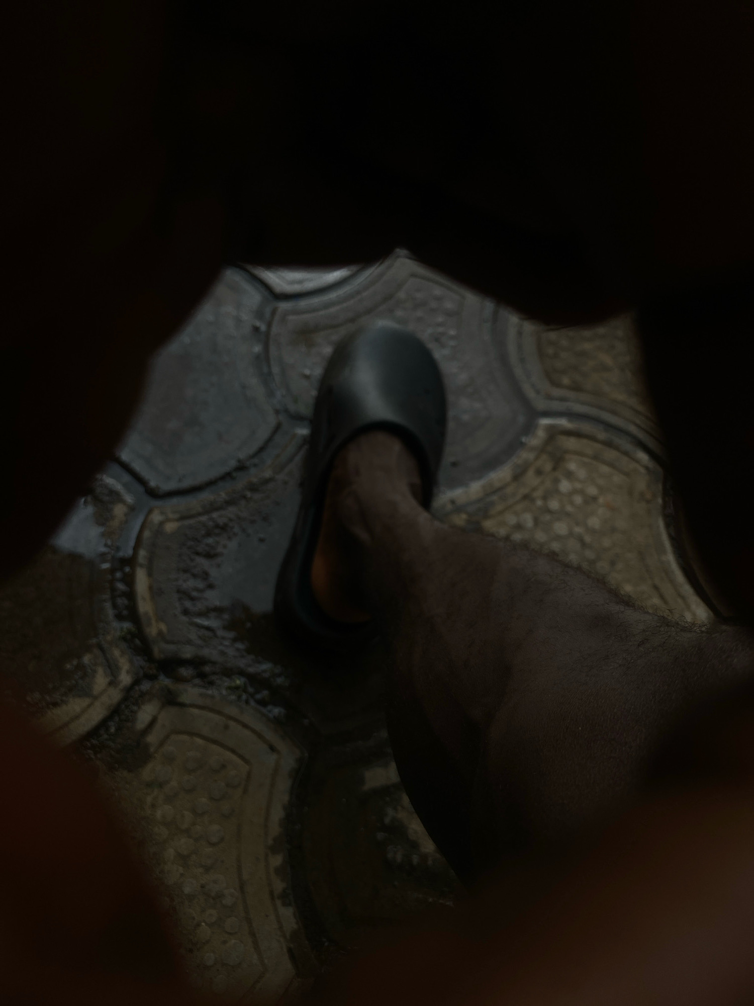A low-angle shot shows a bare leg and foot in a black slide sandal, standing on wet, patterned paving stones. The scene is dimly lit and surrounded by darkness, suggesting it may be an indoor space with poor lighting or nighttime. The paving stones appear to be dark gray and have an octagonal shape with textured inlays. Some areas of the stones are wet, indicating recent rain or cleaning. The leg is hairy and appears to be a darker skin tone.