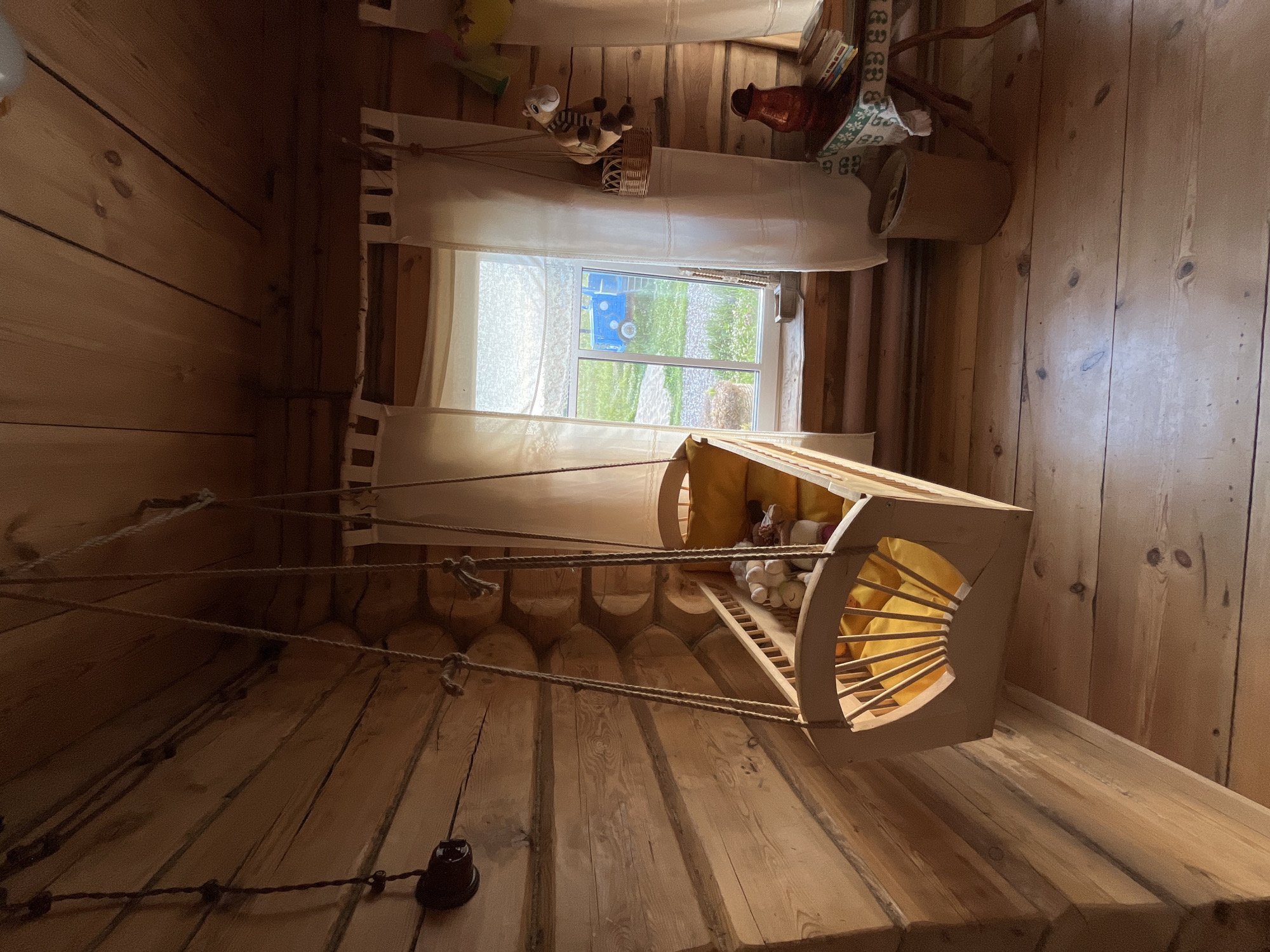 This image depicts a rustic, wooden room in Vybolovo, Russia, likely a bedroom or nursery. A wooden crib, suspended by ropes from the ceiling, hangs in the center of the room. The crib contains several stuffed animals and a yellow mattress. To the right of the crib is a window with white curtains, revealing a green outdoor scene with a blue vehicle visible in the distance. A small wooden table with a red pitcher and books stands to the right of the window. The walls and floor are constructed from natural wood, contributing to the room's cozy, traditional aesthetic. There are no people present in the image.