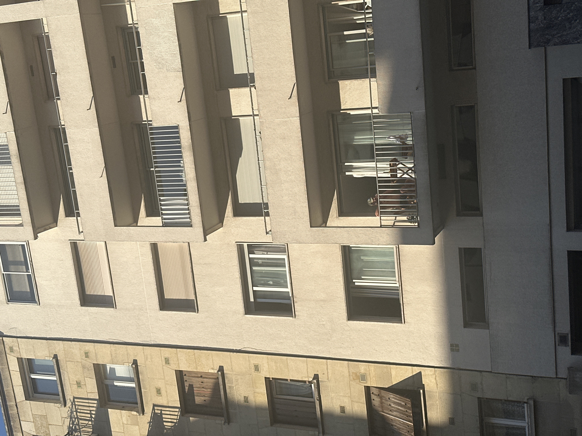 The image shows a close-up, exterior view of a residential building. The building is constructed with two distinct facade materials: the left side features large, light-colored stone blocks, while the right side is made of a light-colored, textured material. Numerous windows and balconies are visible across the different levels of the building. The sun casts long shadows, indicating it is likely late afternoon or early morning. On the lower right balcony, two individuals are visible, one appearing to be sitting at a table. This image does not provide any specific details that would allow for pinpointing its location within Paris, France.