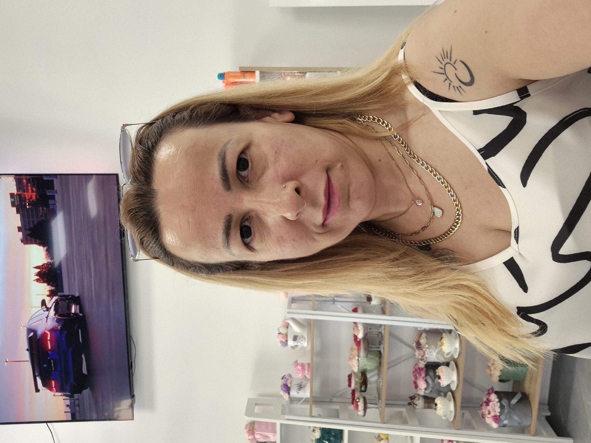 A woman with blonde hair and fair skin is looking at the camera. She is wearing a white tank top with a black abstract pattern and several gold necklaces. She has a tattoo on her right shoulder. In the background, shelves are filled with decorative items, possibly cupcakes or flowers. A television screen on the wall displays an image of a blue car. The setting appears to be an indoor space, potentially a shop.