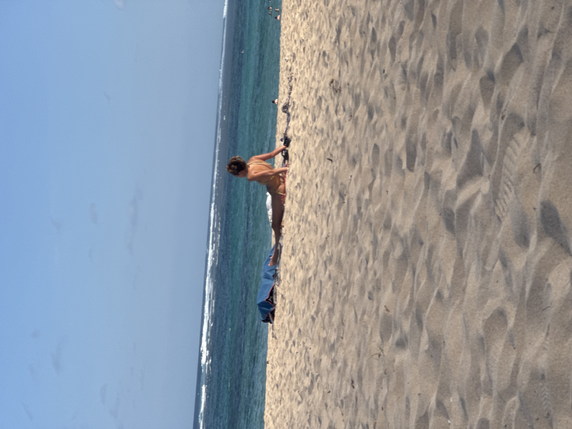 A lone woman in a bikini is sunbathing on a sandy beach in Kuta Selatan, Indonesia. The woman is lying on her stomach with her back to the camera, facing the ocean. The ocean is a deep blue with white waves breaking near the shore. A blue beach umbrella is set up on the sand behind the woman. The sand is light brown and has ripples from the wind. The sky is clear and blue with a few small clouds. There are other people in the water in the distance.