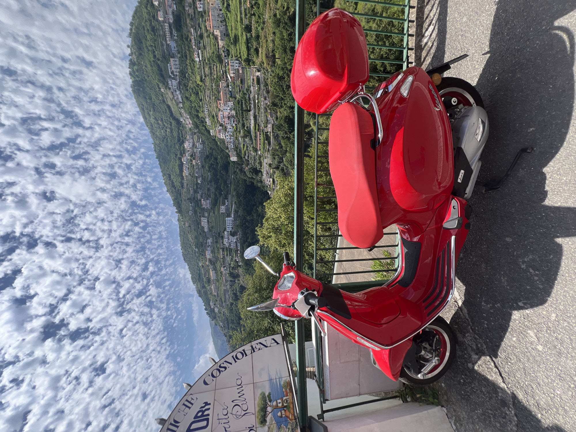 A red Vespa scooter is parked on a paved surface with a green metal railing in the foreground. The scooter faces to the left of the frame and has a red seat and a red top case. Behind the scooter, a steep hillside covered in greenery and dotted with white buildings rises into the distance. The sky is bright blue with numerous small, white clouds. To the left of the scooter, a white tiled circular sign with blue lettering is partially visible. The image appears to be taken on a sunny day, possibly in Ravello, Italy, given the architecture and landscape typical of the Amalfi Coast. No people are visible in the image.