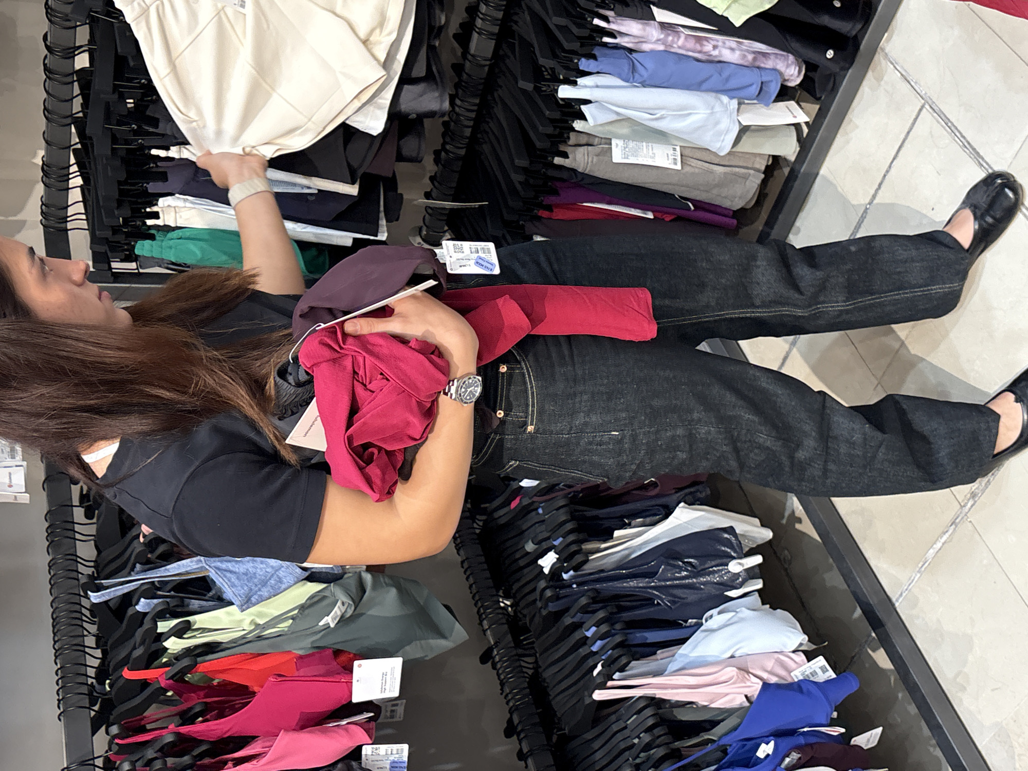 An overhead shot captures a person shopping in a retail environment, identified as a Lululemon athletic wear store based on visible tags, in San Francisco Ocotlán, Mexico. The individual, with long brown hair, is wearing a black short-sleeved top, dark denim wide-leg jeans, and black flat shoes, with a silver-toned watch on their left wrist.
The person is actively browsing, with their right hand reaching into a clothing rack displaying cream-colored garments, while their left hand holds several items in vibrant shades of red and purple. The store features multiple black metal clothing racks arranged in aisles, filled with a diverse range of activewear items. These garments are neatly hung on black hangers or folded on shelves, showcasing a varied palette including black, green, light blue, grey, pink, dark blue, purple, and red.
Numerous white tags are visible on the clothing, many clearly displaying the "lululemon" brand logo. One tag held by the shopper explicitly shows "lululemon," "RETAIL PRICE," and "MXN" (Mexican Peso), with a partially visible price of approximately "8745 MXN." The store's interior features a light-colored tiled floor. The scene depicts a focused shopping moment in an indoor setting, likely under artificial lighting.