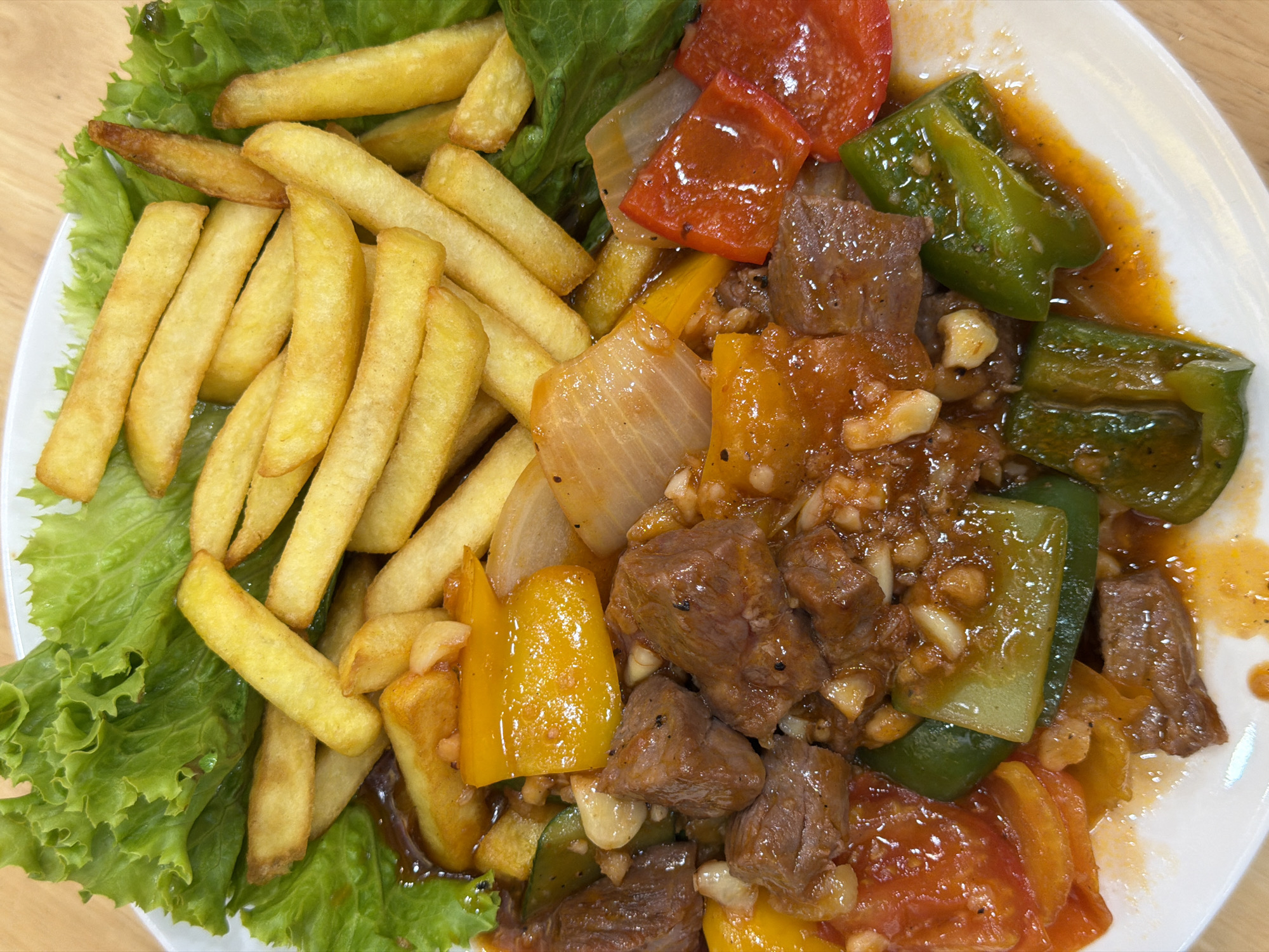 The image shows a plate of food with a serving of french fries and a meat and pepper stir-fry. The french fries are piled on a bed of lettuce on the left side of the plate. The stir-fry consists of chunks of meat (likely beef), green, red, and yellow bell peppers, and onions in a glossy sauce. The dish appears to be a combination of Western and Asian cuisine elements. The plate is white and set against a light brown wooden surface. The location is Nha Trang Ward, Vietnam.