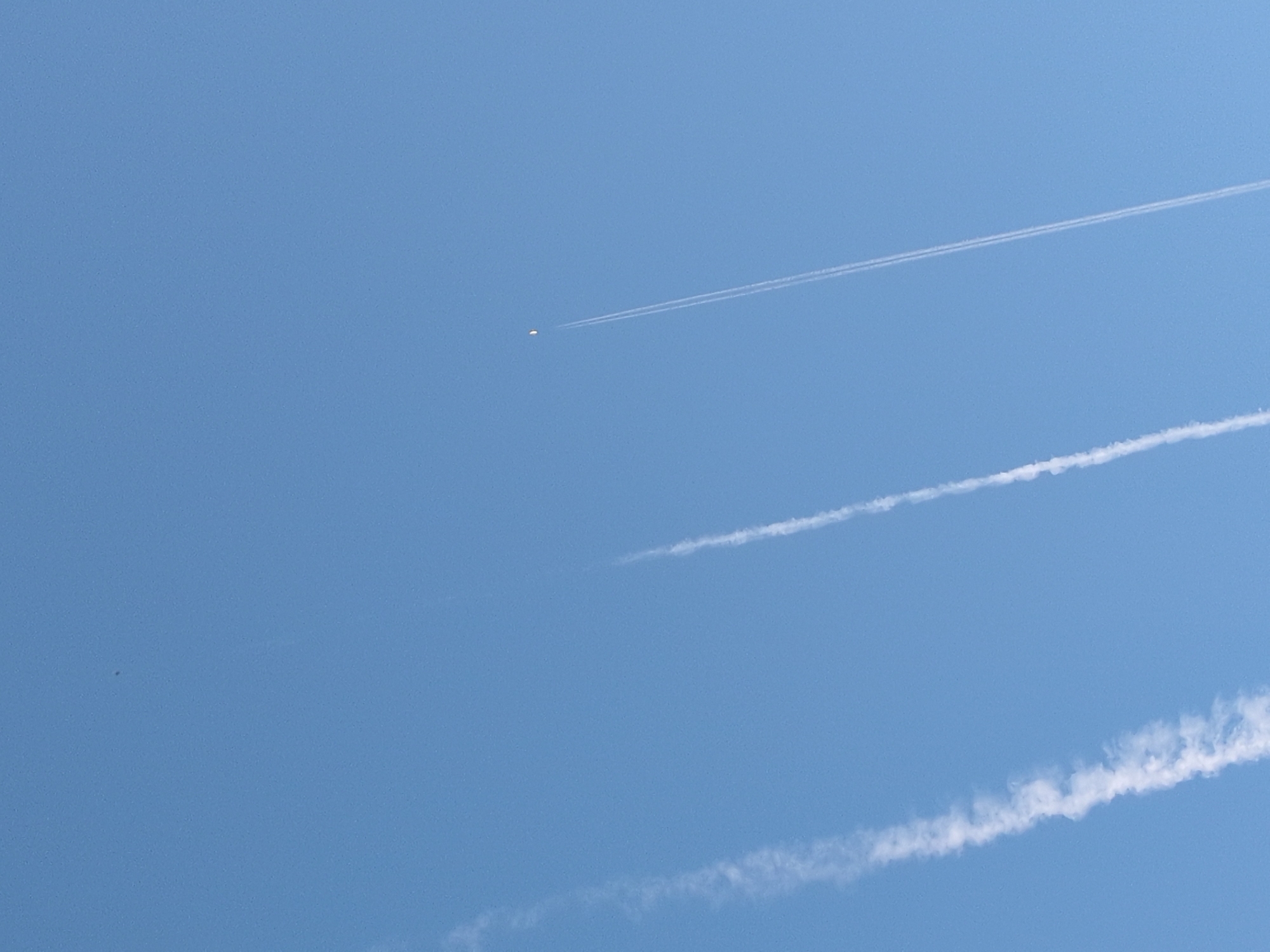 The image displays a clear, light blue daytime sky. Three distinct white linear contrails from high-altitude aircraft are visible. The leftmost contrail is broad and appears to be dispersing. A thinner, more defined contrail runs diagonally upwards in the mid-left portion of the frame. The rightmost contrail is narrow, sharp, and extends upwards towards the top-right. In the upper right quadrant, a bright, yellowish-white crescent-shaped celestial body, consistent with the daytime Moon, is prominently featured above the sharpest contrail. A very small, indistinct dark speck is located in the upper left quadrant. No human subjects, ground features, or specific landmarks are visible. The image itself provides no geographical markers to confirm its capture in Sochi, Russia.