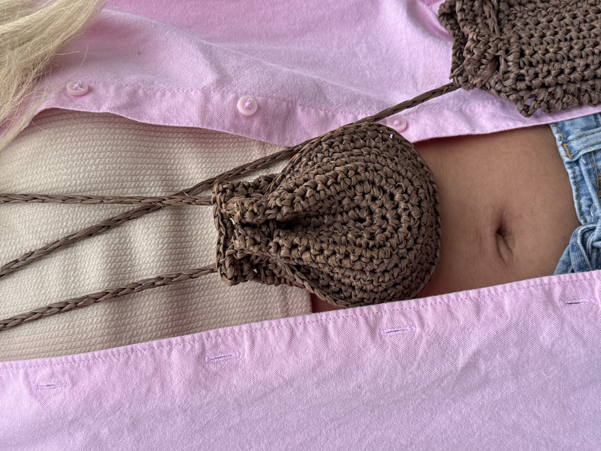 The image is a close-up shot of a person wearing a pink button-up shirt over a beige ribbed tank top. The person has a small, brown, woven drawstring bag hanging around their neck, and another similar bag is partially visible in the lower right corner. The person's midriff is exposed, showing their navel. The background is a plain pink fabric. There is no indication of the city of Burgas, Bulgaria, or any other specific location in the image.
