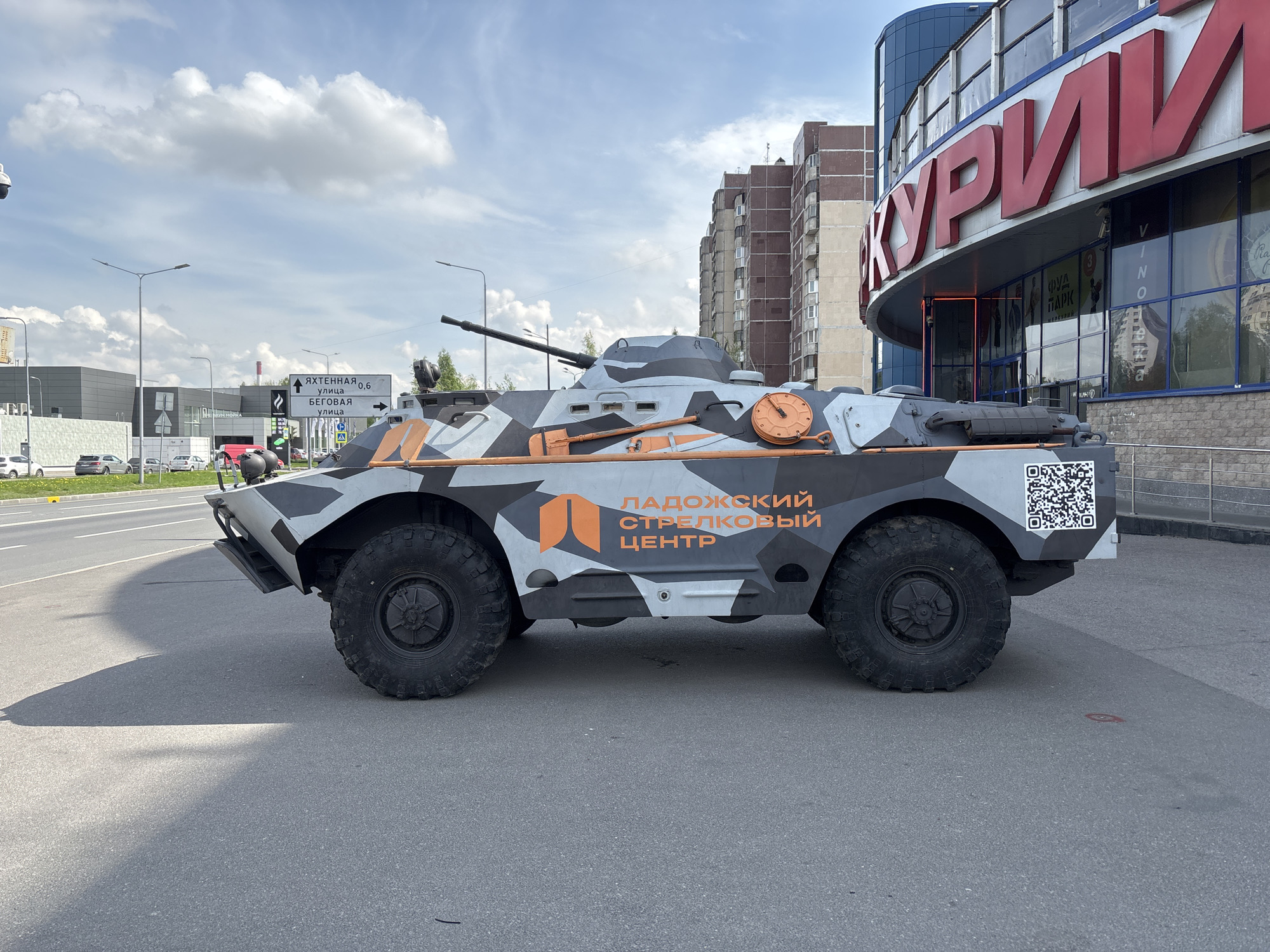 The image shows an armored personnel carrier (APC) parked on a paved surface in what appears to be an urban setting in Saint Petersburg, Russia. The APC is painted in a camouflage pattern of gray, black, and orange. The text "ЛАДОЖСКИЙ СТРЕЛКОВЫЙ ЦЕНТР" (Ladoga Shooting Center) is printed on the side, along with a logo. A QR code is located at the rear of the vehicle.
Behind the APC is a building with a large red sign that reads "КУРИИ". The building has glass windows and a curved façade. Further back, there is a multi-story residential building with balconies. A street sign pointing to "ЯХТЕННАЯ" (Yachtennaya) street and "БЕГОВАЯ" (Begovaya) street can be seen in the background. Several light poles and other commercial buildings are also visible, and the sky is partly cloudy.