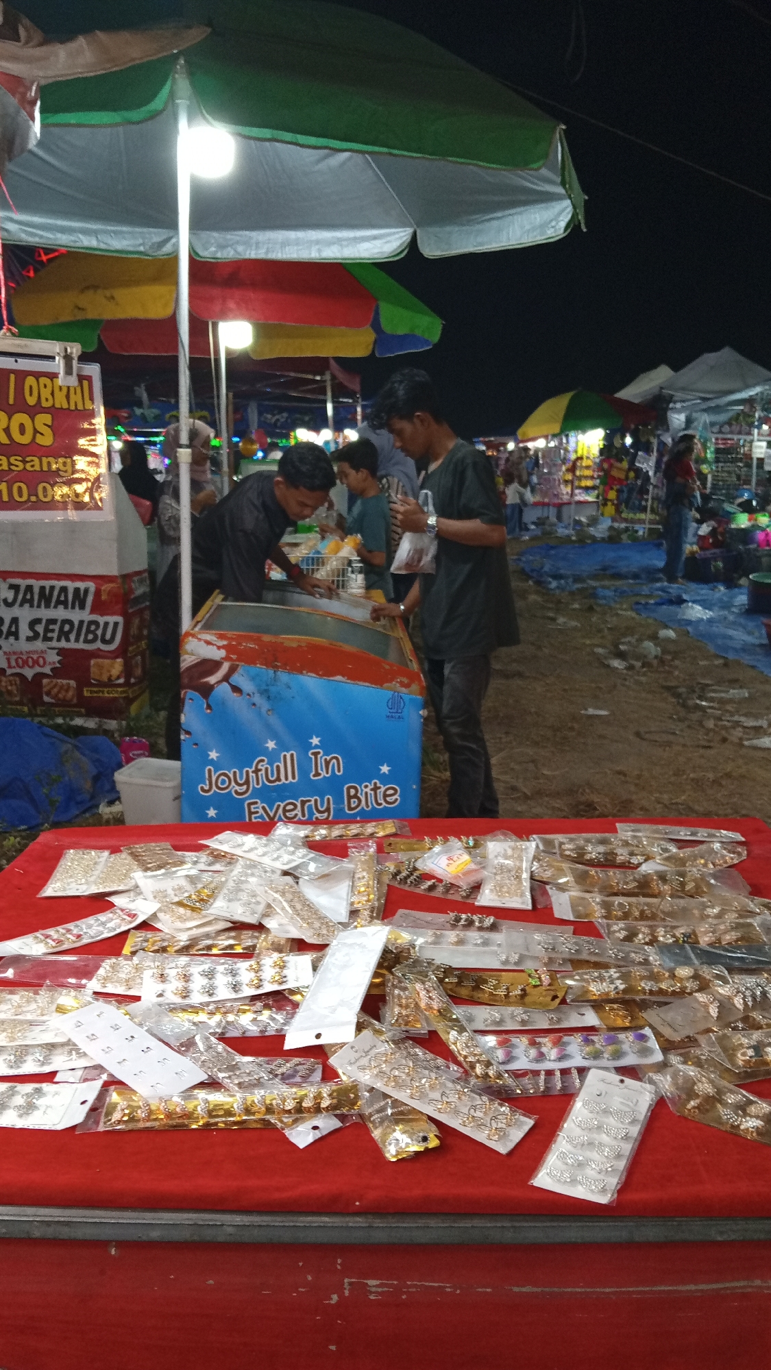 The image captures a night market scene in Blangcotbaroh, Indonesia. In the foreground, a red-covered table is laden with numerous small, individually packaged accessories, primarily jewelry such as earrings and hair clips, displayed in clear plastic packaging. Many of these packages are gold or silver in color, and some feature decorative elements like butterflies. Behind the table, an open-top cooler is visible, emblazoned with the text "Joyfull In Every Bite." Three young men are positioned around the cooler. One, wearing a black shirt, appears to be reaching into the cooler. Another, in a dark t-shirt and black pants, is holding a plastic bag. The third young man, in a blue t-shirt, is also near the cooler.
Overhead, colorful umbrellas – green, yellow, and red – provide shade or decoration for the stalls. The background reveals more stalls with merchandise and people browsing, set against a dark, night sky. The ground appears to be dirt or sand, covered in places by blue tarps. The overall atmosphere suggests a bustling marketplace.