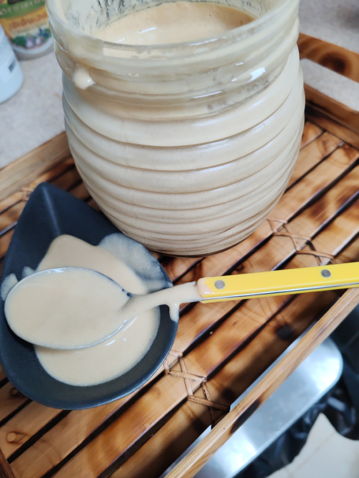 A close-up, high-angle, static shot shows a jar filled with a light brown, viscous liquid. The jar is made of clear glass with horizontal ridges and is placed on a wooden tray. A dark grey, leaf-shaped dish containing some of the same liquid sits in front of the jar. A spoon, with a yellow handle, is partially submerged in the dish, scooping up the liquid. The background is blurred, but appears to be an indoor setting with some kitchen items visible. There is no indication of the city of Dubai or the United Arab Emirates in the image.