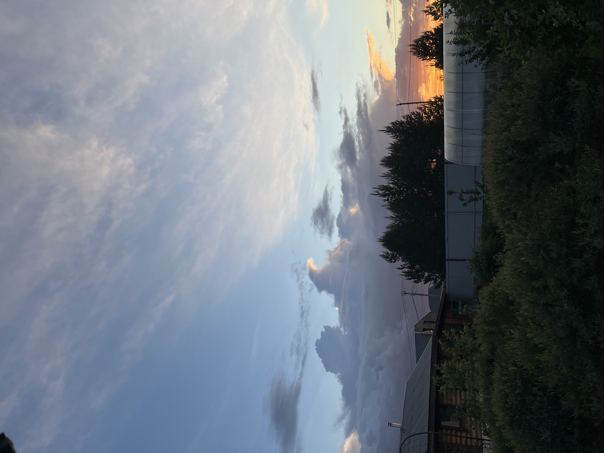 The image shows a sky filled with a mix of grey and white clouds, with a patch of orange and yellow sky visible near the top of the frame. In the lower right of the image, there are trees and a building. The location is suggested to be Chaikovsky, Russia.