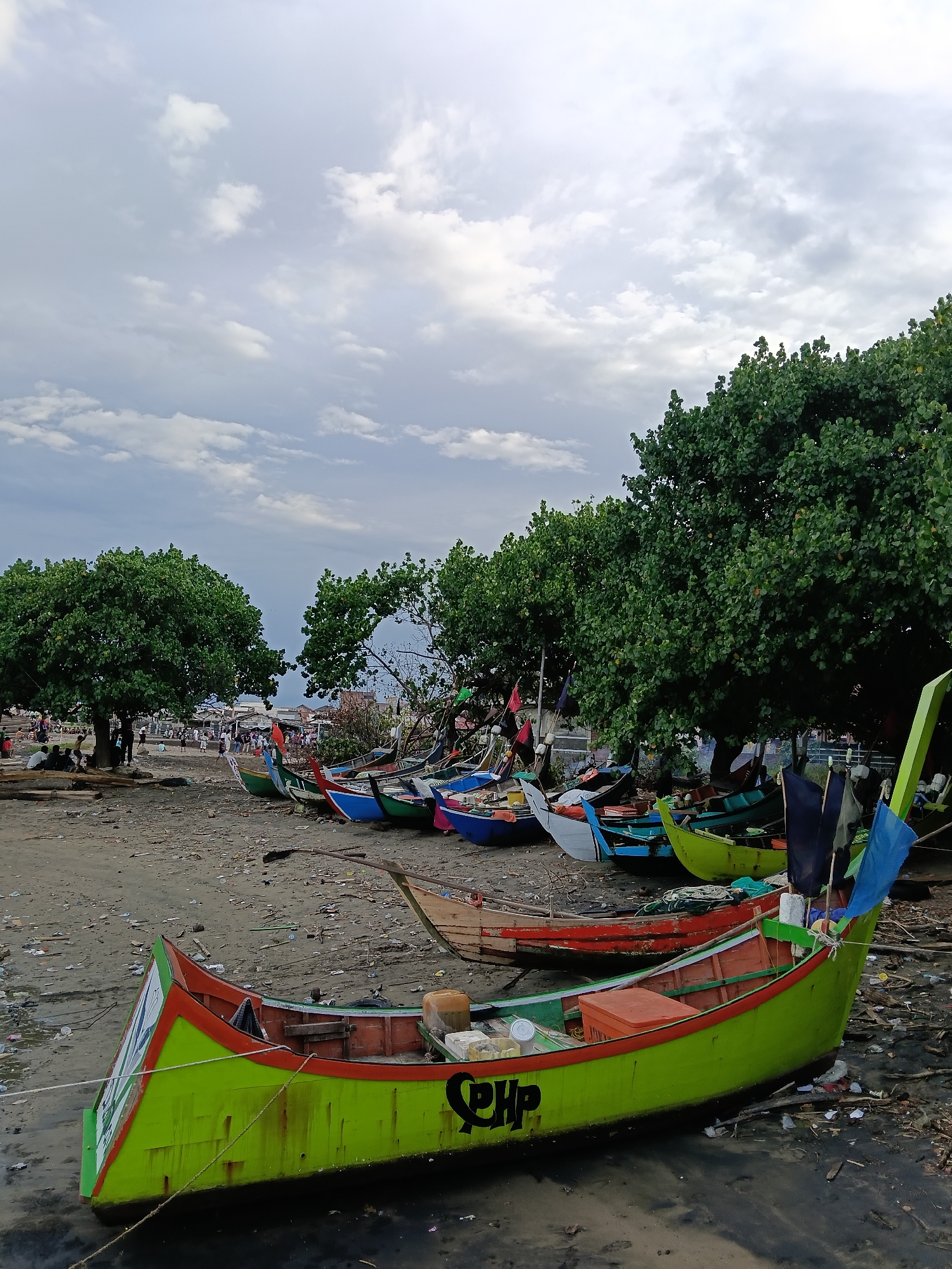 The image shows a coastal scene in Lhokseumawe, Indonesia. Several colorful fishing boats are pulled up on a beach littered with trash. The boats are of various sizes and colors, primarily green, blue, and red. Some of the boats have flags or banners attached. A line of trees with dense green foliage runs along the back of the beach. In the background, indistinct figures of people can be seen moving about. The sky is overcast with grey and white clouds. The beach itself is dark and muddy.