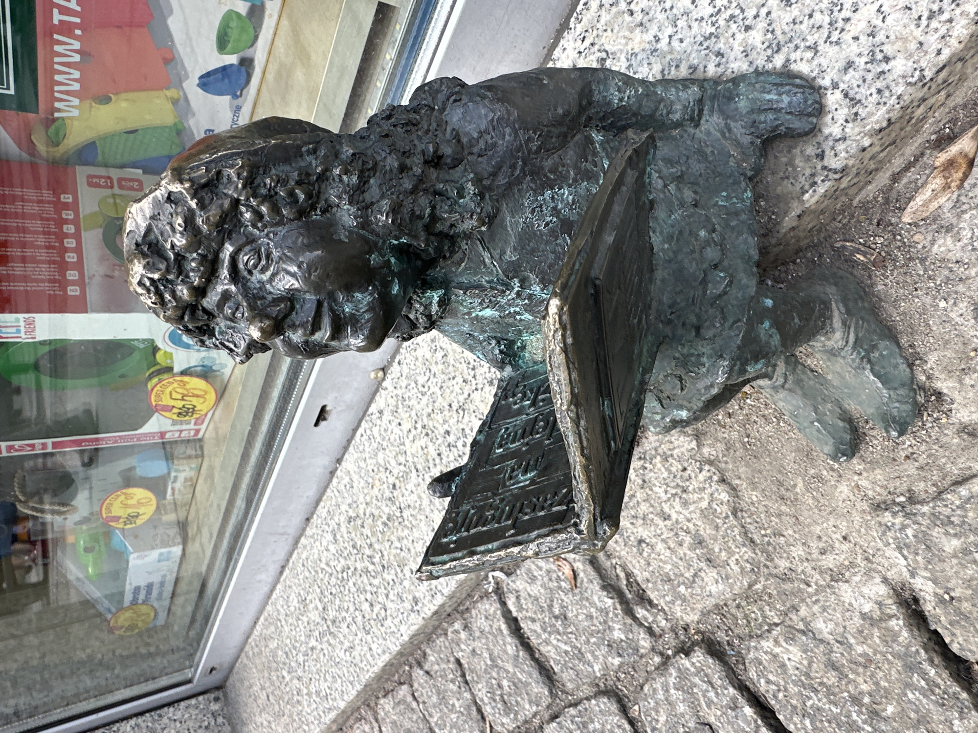 A bronze dwarf statue sits on the cobblestone sidewalk next to a building in Wrocław, Poland. The statue, likely a gnome or mythological figure, appears to be reading a book, as it is hunched over with a rectangular object in front of it with inscribed text. The statue has a heavily textured surface, reflecting its age and material. The building's storefront is visible behind the statue with sale stickers on the glass of a shop. The lighting suggests a bright, overcast day.