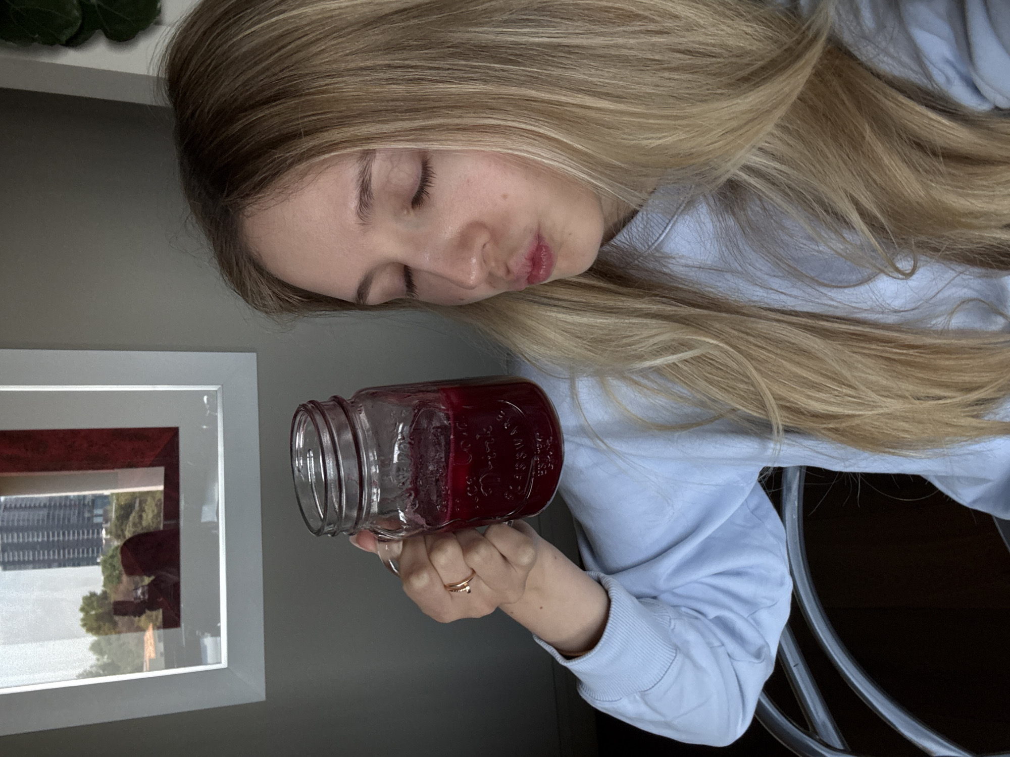 A fair-skinned, blonde woman with closed eyes is holding a mason jar filled with a reddish-purple liquid. She's wearing a light blue long-sleeved sweatshirt. Her blonde hair is long and loose. She is wearing a ring on her left ring finger. Behind her to the left is a framed picture with a view of the city of Mexico City, Mexico visible through the window. The surrounding environment is a neutral-toned wall and a dark floor. The exact location in Mexico City, Mexico cannot be determined from the image.