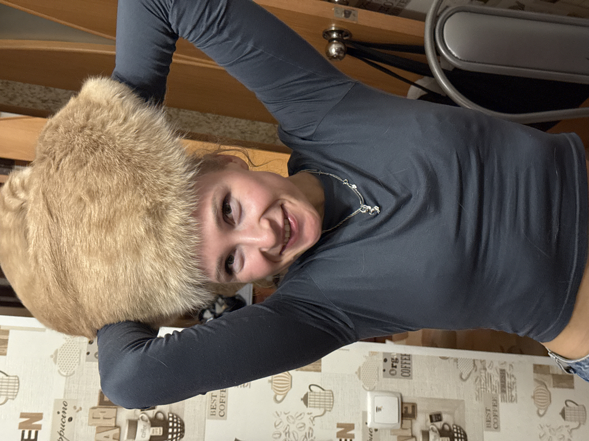 A young woman wearing a gray long-sleeved shirt and a silver necklace is holding a large, furry, light brown object near her head, possibly a hat or animal. She is smiling at the camera. The background consists of wooden elements, possibly furniture or a wall, a gray metal object, and a kitchen counter or table covered with a patterned tablecloth featuring images of coffee pots and coffee-related text. The location is potentially in Perm, Russia, based on possible regional context.