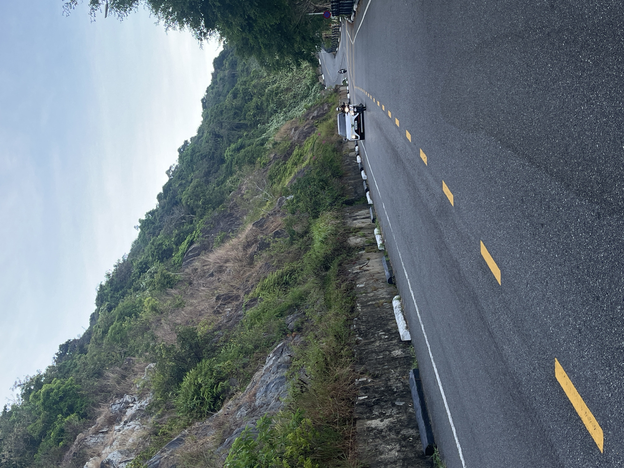 The image shows a road winding along a mountainside. On the road, a white car and at least one motorcycle are traveling. The road has yellow dashed lines and a white shoulder line. A low concrete barrier runs alongside the road. The mountainside is covered in vegetation. The sky is overcast. The location is likely on the Hải Vân Pass in Đà Nẵng, Vietnam, as this is a common scenic mountain road with similar features.