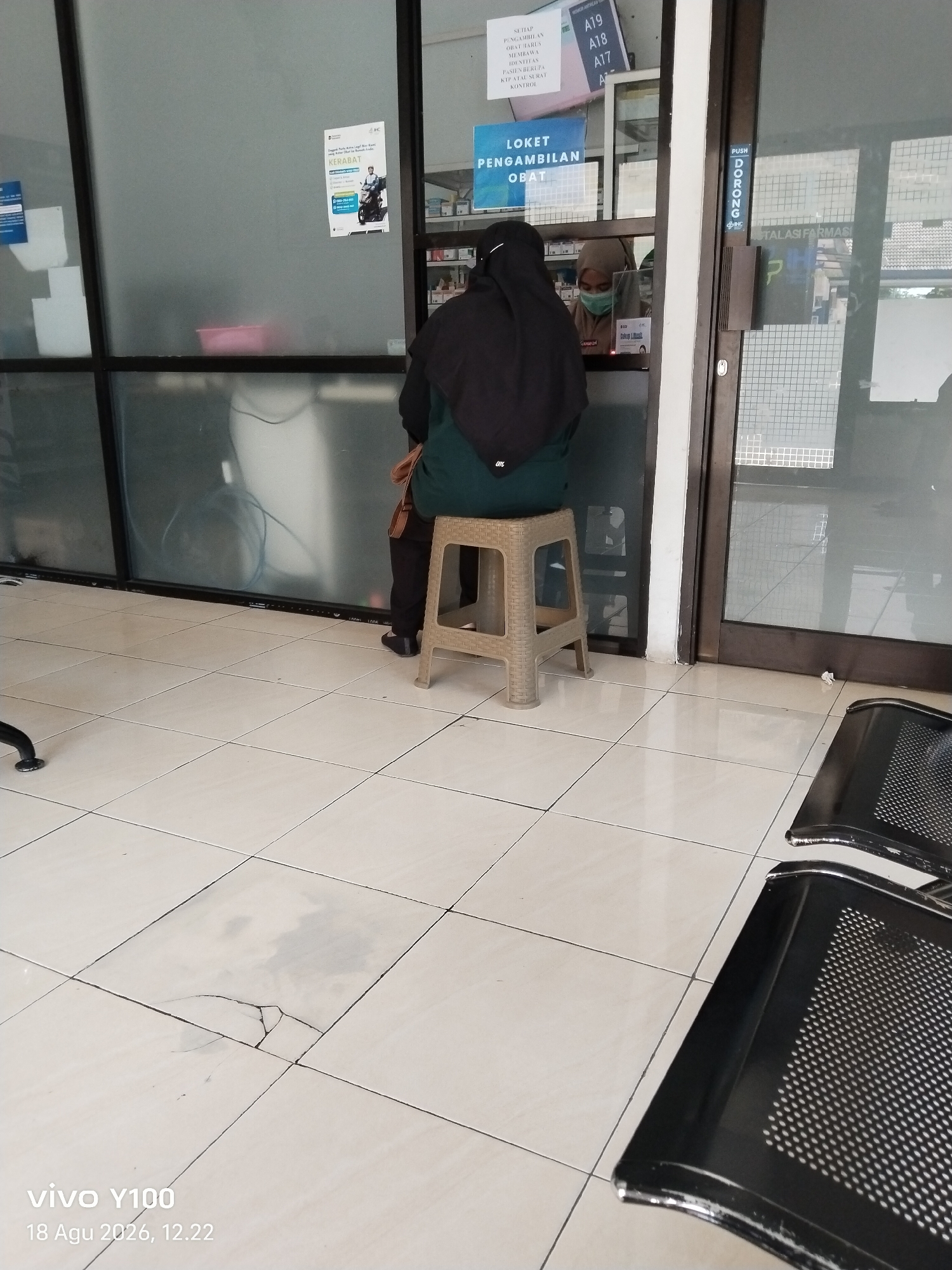 The image shows the interior of a facility, possibly a clinic or hospital pharmacy, with a tiled floor and large glass windows. In the foreground, there are two black metal chairs with perforated seats. A woman wearing a dark hijab and a green garment is seated on a light-colored plastic stool in front of a glass counter. She is partially obscured by the glass. Behind the counter, another person wearing a light-colored hijab and a face mask is visible, appearing to be interacting with the seated woman. Signs are posted on the glass, including one that reads "LOKET PENGAMBILAN OBAT" (Medicine Collection Counter) and others with alphanumeric labels. The overall environment appears to be a waiting area or service counter. The timestamp "vivo Y100 18 Agu 2026, 12.22" is overlaid on the bottom left of the image, suggesting it was taken on August 18, 2026, at 12:22. The specific location in Patokan, Indonesia, is not explicitly indicated in the visual information, but the text on the signs appears to be in Indonesian.