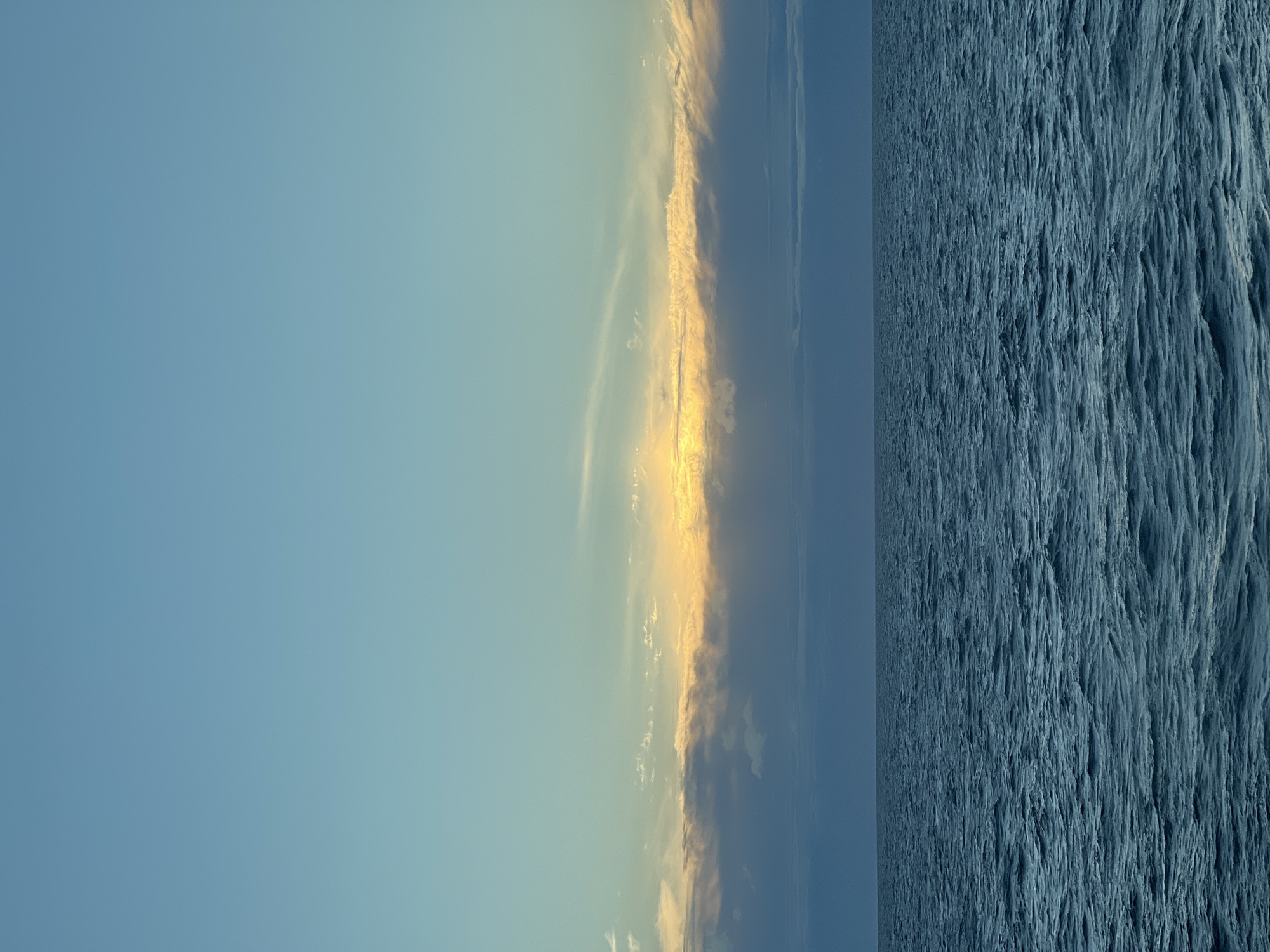 A horizontal image shows a calm sea under a pale blue sky with scattered clouds. The sun is setting, casting a warm orange glow on the horizon. The water is a deep blue with gentle ripples. The horizon line is sharp, dividing the sea and sky. The scene is serene and peaceful.