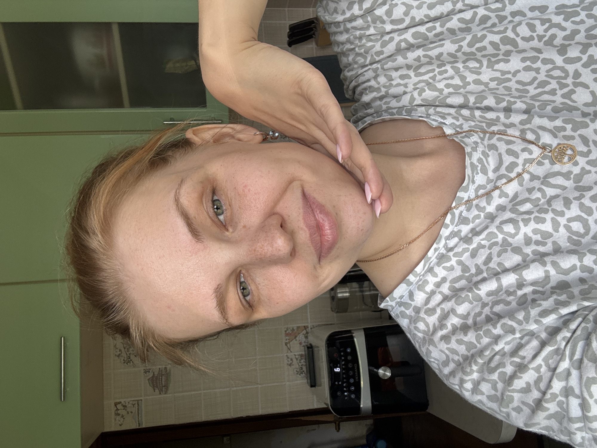 A fair-skinned female, with light-colored hair, green/hazel eyes, and visible freckles on her nose and cheeks, is taking a front-facing selfie within a kitchen in Rusanovo, Russia. She exhibits a gentle smile, gazing directly at the camera. Her left hand, featuring light pink manicured nails, supports her chin. She is wearing a light grey and white leopard-print t-shirt, a gold-colored chain necklace with a circular 'Tree of Life' pendant, and a rose gold-toned earring with a light blue square-cut stone in her visible right ear. The background reveals a kitchen equipped with light green upper cabinets, one displaying a horizontal silver handle, and a tiled backsplash featuring decorative panels with traditional Asian-style landscape imagery. On a light-colored countertop, a black and silver 'DEMIANO' air fryer with a digital display showing '6.' is visible to the subject's left. A clear pitcher or blender is partially discernible next to the air fryer, and a knife block is visible in the far right background.