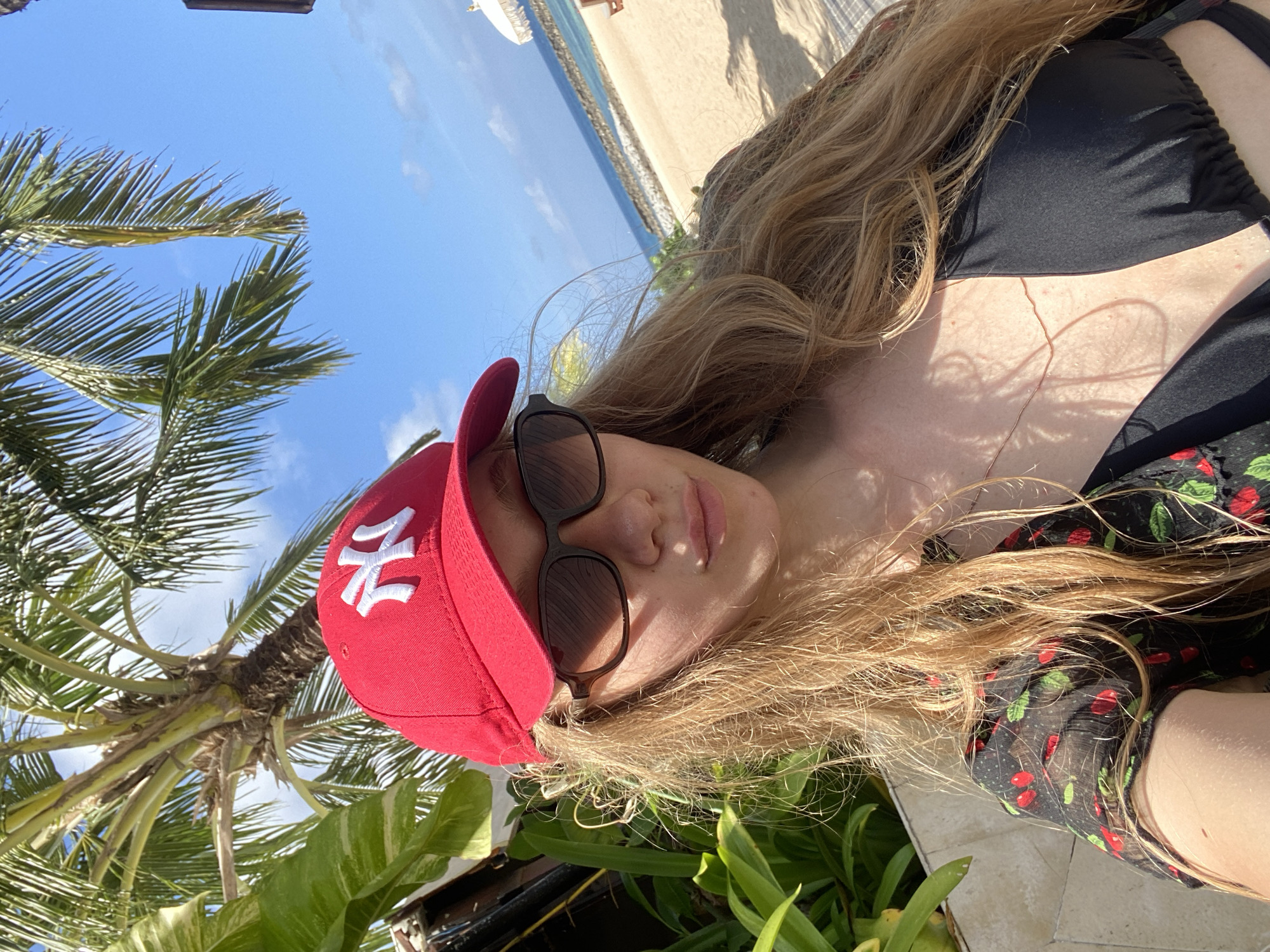 The image shows a woman lying on a tiled surface, presumably outdoors, with a beach and ocean visible in the background. The woman is wearing a black bikini top with a sheer, cherry-printed cover-up, sunglasses, and a red baseball cap with a white logo. A palm tree and various green plants surround her. Based on the vegetation and the beach visible in the background, the location is likely in the coastal area of Kuta Selatan, Indonesia.