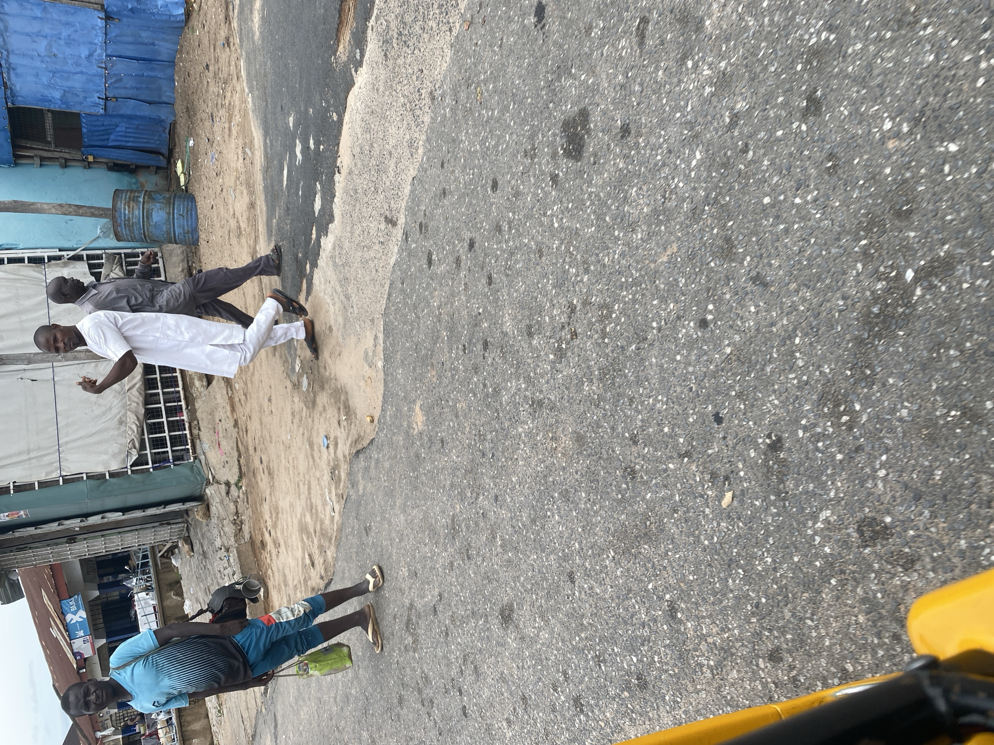 The media file depicts a street scene in Jalingo, Nigeria. In the foreground, a person is walking towards the camera, wearing a blue t-shirt and blue shorts, with flip-flops on their feet and a green bag slung over their shoulder. Behind them, to the left, two men are walking away from the camera. One is wearing a white traditional outfit, and the other is wearing a gray shirt and dark pants. They are walking on a dirt and asphalt road.
To the left of the road, there are buildings with blue and light blue walls, some covered with corrugated metal. A large blue barrel is visible near one of the buildings. Further down the street, more buildings with various signs are visible. The time of day appears to be daytime, and the weather seems to be overcast or hazy. The overall scene suggests a typical bustling street in a Nigerian town, with people going about their daily activities. There is no visible text in the image that can be clearly read.