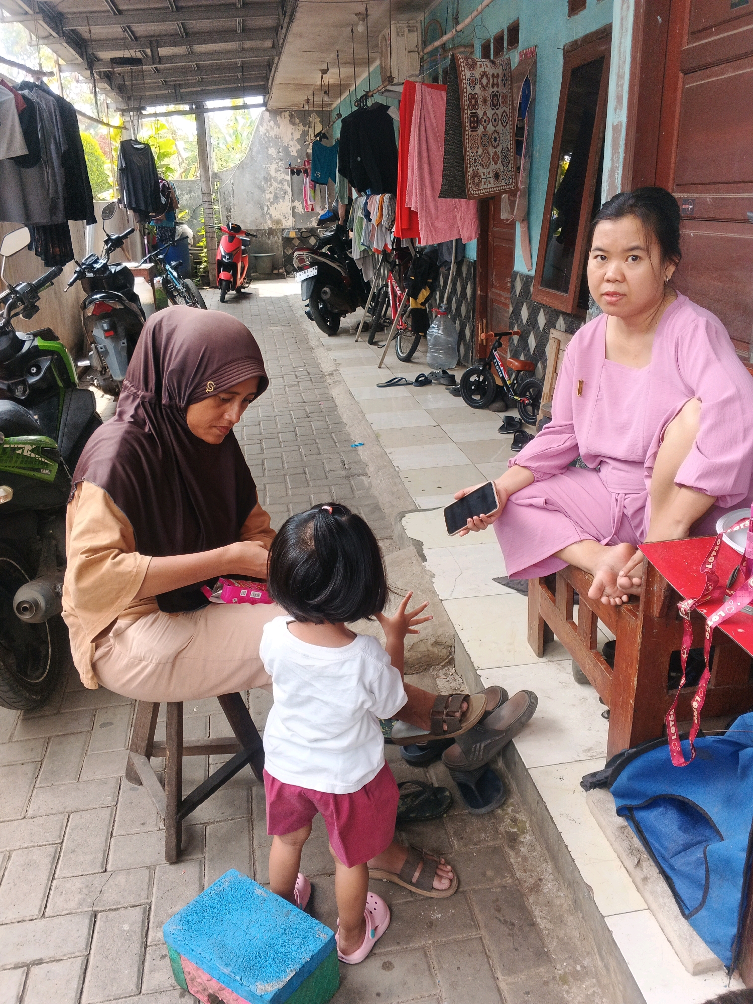 In the city of Kadu, Indonesia, an outdoor scene unfolds under a covered walkway. Three individuals are present: a woman wearing a pink outfit is seated on a wooden stool on the right, holding a smartphone. To her left, another woman in a brown hijab and tan clothing is also seated on a wooden stool, facing a young child. The child, wearing a white t-shirt and pink shorts, stands in front of the woman in the hijab. The environment is paved with gray rectangular bricks and features several motorcycles parked along the side. Clothes are hung on a line overhead. The background shows a building with turquoise walls and wooden doors. A small red and brown toy bicycle and various pairs of sandals are scattered on the ground. A blue bag rests near the woman in pink.