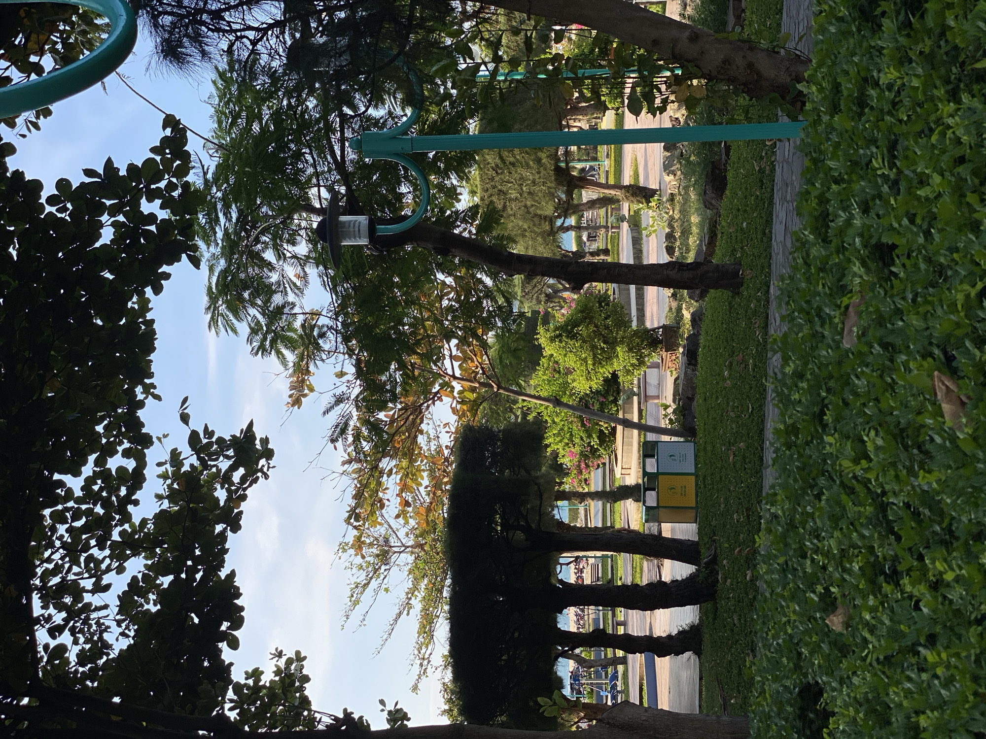 A park scene is depicted in Nha Trang Ward, Vietnam, under a bright sky with scattered clouds. In the foreground, lush green bushes fill the lower portion of the frame. A paved pathway winds through the park, leading towards the background.
To the left of the pathway, several trees with dark, thick trunks stand, one of which is partially obscured by the foreground bushes. Beyond these trees, more trees with rounded canopies are visible, and a row of benches and playground equipment is set against the backdrop of a blue ocean and sandy beach.
To the right of the pathway, a teal lamppost with a decorative curved top stands out. Behind the lamppost, a variety of trees and bushes are present, some with green foliage and others with hints of autumnal colors. A small, green, flowering bush is also visible.
In the middle ground, between the trees and the beach, are two trash receptacles, one yellow and one blue, indicating a public area. The ground is covered in a mix of grass and small, low-lying green plants. The overall impression is a well-maintained park adjacent to a beach, offering recreational and scenic opportunities.