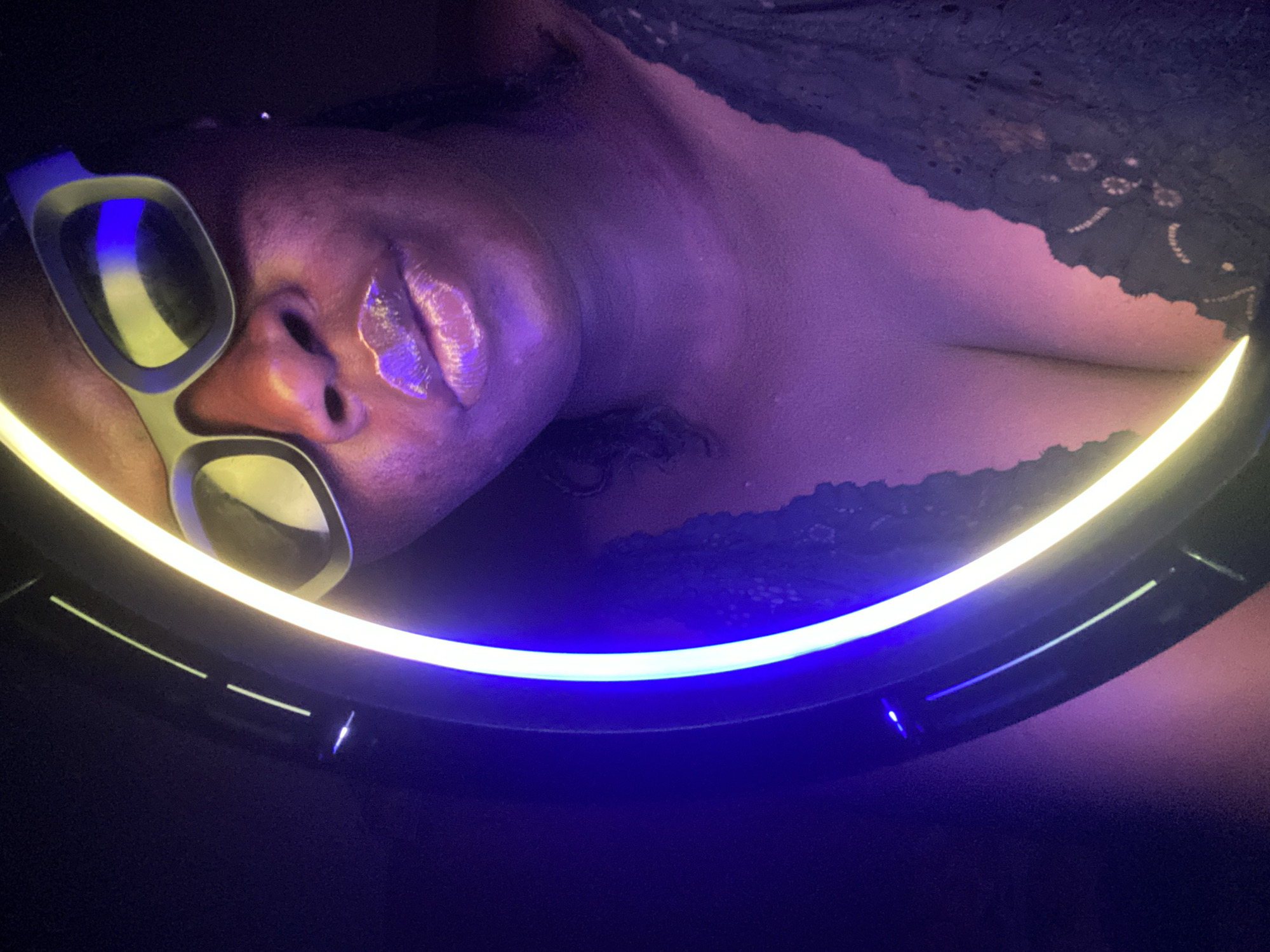 A close-up, angled shot features a person with brown skin wearing light-rimmed glasses with white and gray accents on the face. The subject's lips are glossy and a light-colored substance appears around the right nostril. The person is also wearing a lacy black top. A circular LED light is positioned around the subject, casting a bright yellow light. The scene is predominantly purple and dark blue. There is no indication this is in Maigizo, Nigeria.