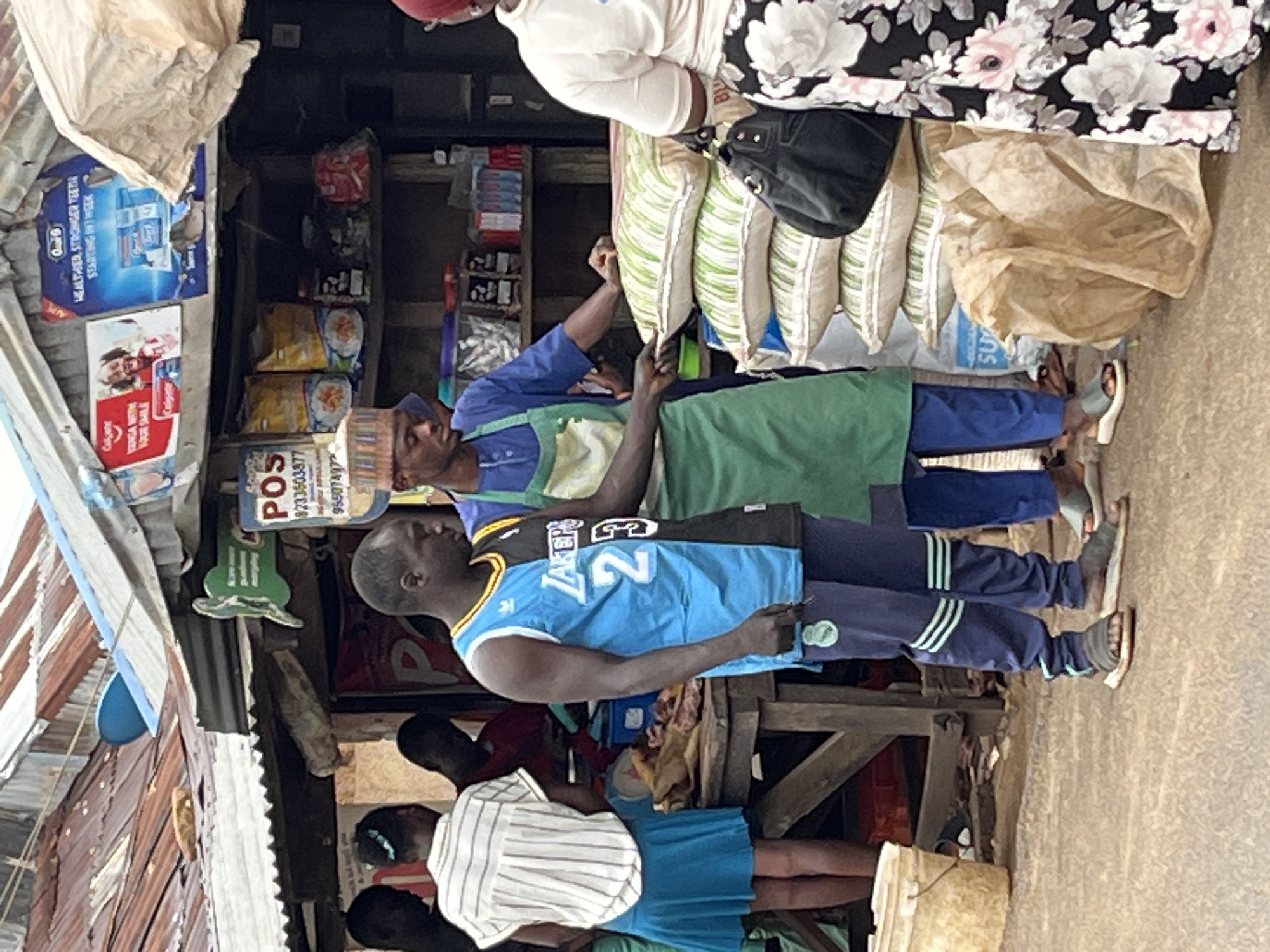 The image shows a street scene in Jos, Nigeria. A group of people are gathered around a small store. A man wearing a Lakers jersey with the number 2 is standing in front. A man in a blue shirt and green apron is behind him, presumably the storekeeper. A POS sign is visible above the store. Sacks are seen at the back of the store, and various items are displayed on shelves. The environment is a busy street market with corrugated iron roofing.