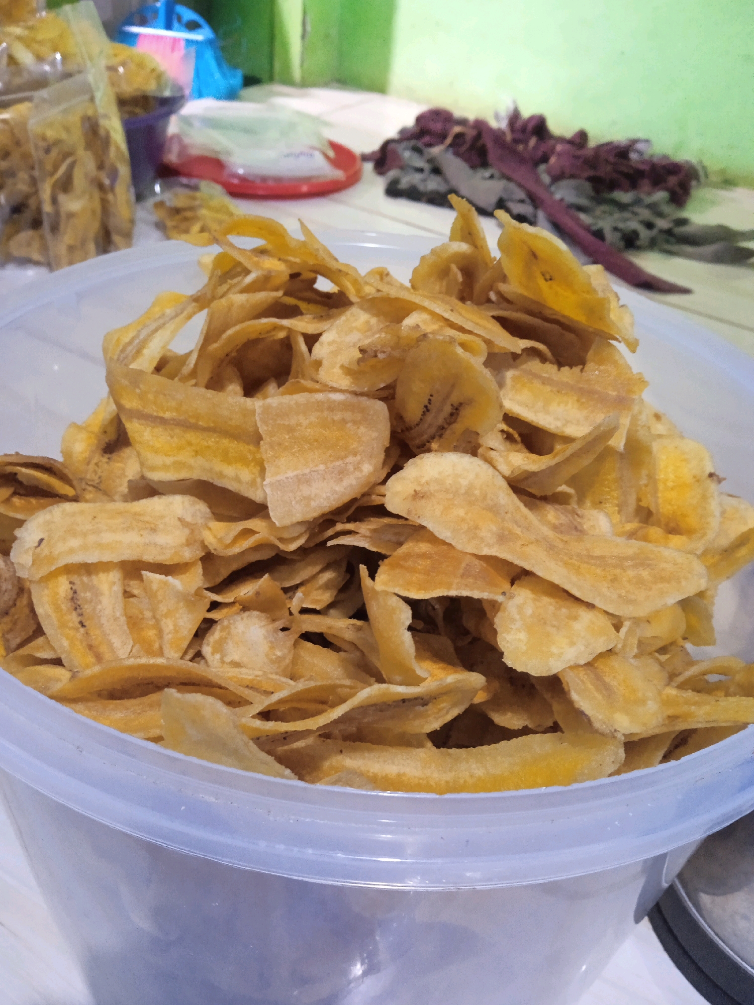 This image, likely taken in Banjarmasin, Indonesia, shows a close-up of a large, clear plastic container overflowing with banana chips. The banana chips are thin, irregularly shaped, and a light golden-yellow color, with some darker brown specks indicating ripeness or slight caramelization. They are piled high, filling most of the container, with some spilling over the rim.
In the background, slightly out of focus, are several plastic bags also containing banana chips, suggesting a small-scale production or storage of these snacks. A red plate with white tissue paper sits in the mid-ground, and behind it, a pile of what appears to be dried leaves or fabric in shades of red and gray is visible. The overall setting suggests a casual, possibly domestic, environment where food is being prepared or stored. The lighting is natural and appears to be from an overhead source, creating soft shadows.