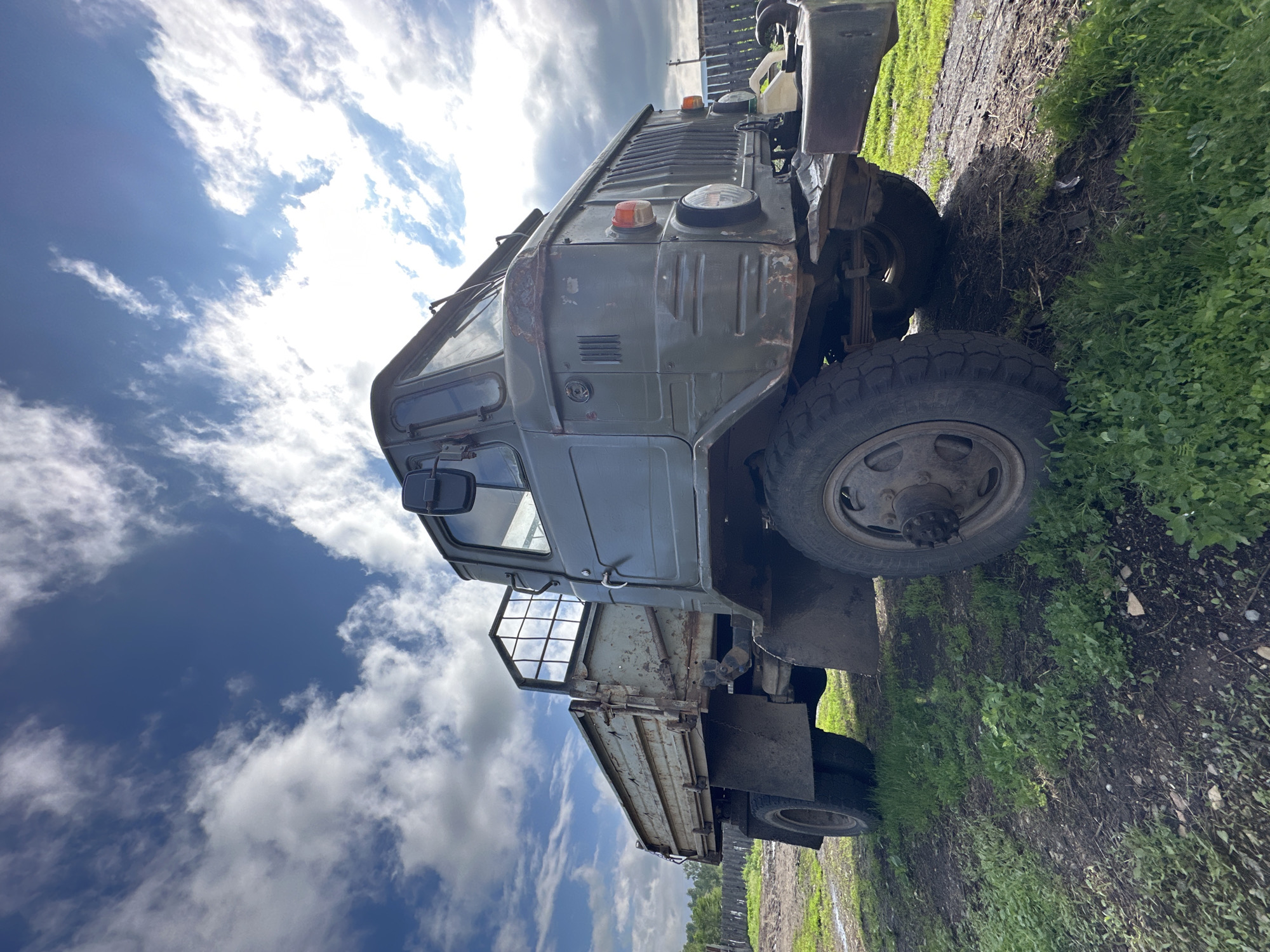 An old, olive-drab, four-wheeled truck is parked on a dirt and grass area. The truck is seen from a low angle, with the sky and clouds above it. The truck has a flatbed cargo area with a metal cage at the back. The driver's side of the truck is visible, showing the door, a side mirror, and the front fender. The front of the truck has a large grille and round headlights. The truck appears weathered and slightly rusted. In the background, there is a wooden fence and some trees. The ground around the truck is covered in patches of green grass and bare dirt. The sky is blue with scattered white clouds.